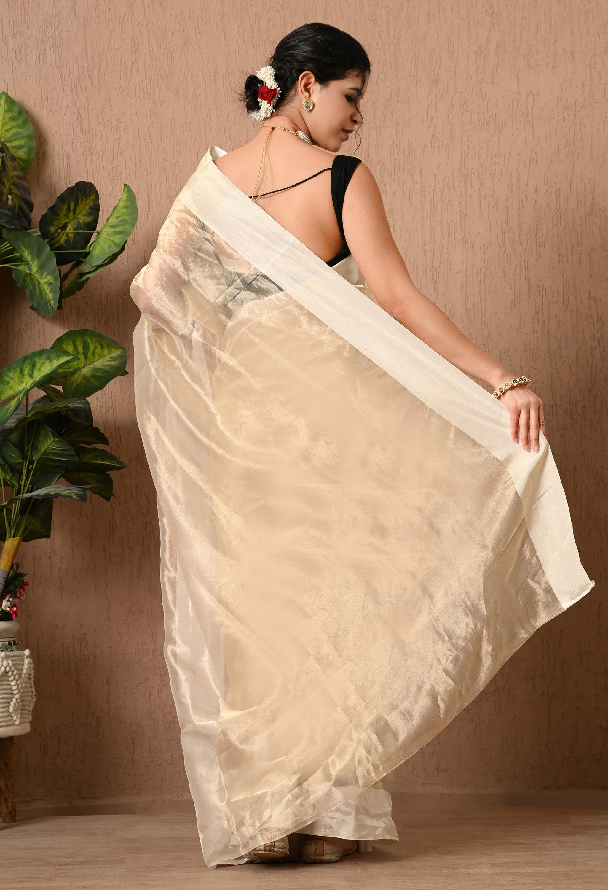 A back view of a woman showcasing the drape of a light golden handwoven tissue saree from The Weavers Shop by Raavi. The image highlights the shimmering, sheer fabric and the distinct cream-colored satin border running along the edge. She is wearing a black sleeveless blouse, with her hair styled in a low bun adorned with white and red flowers. She stands against a neutral textured wall next to a large green indoor plant.