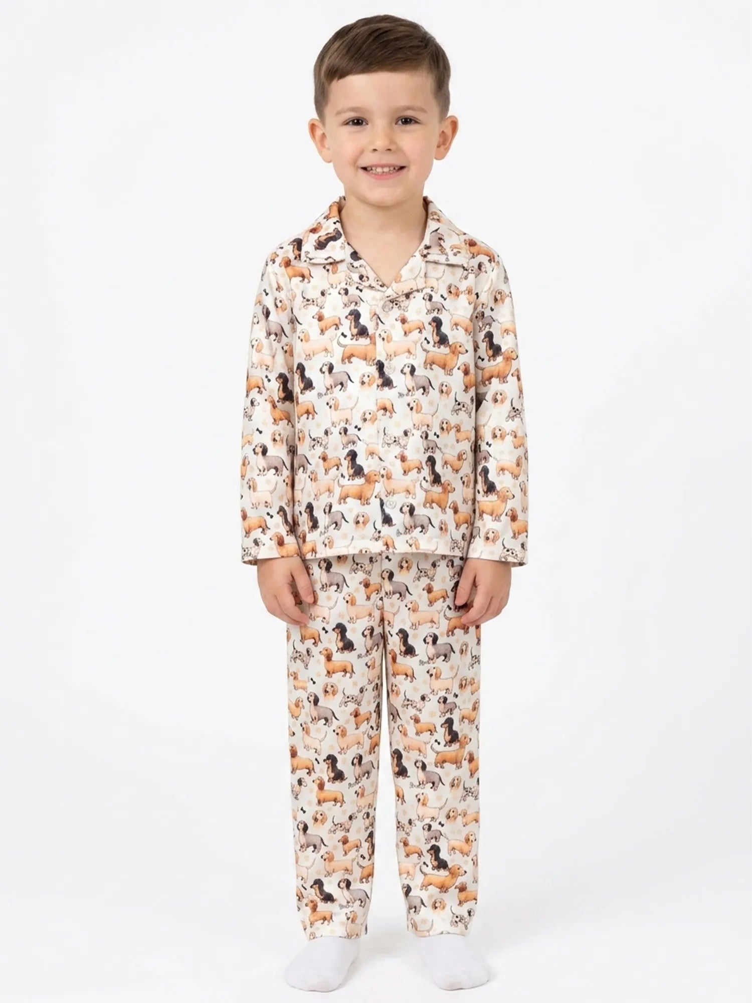 A young boy standing against a plain white background wearing a cream-colored satin pajama set with an all-over print of dachshund dogs.
