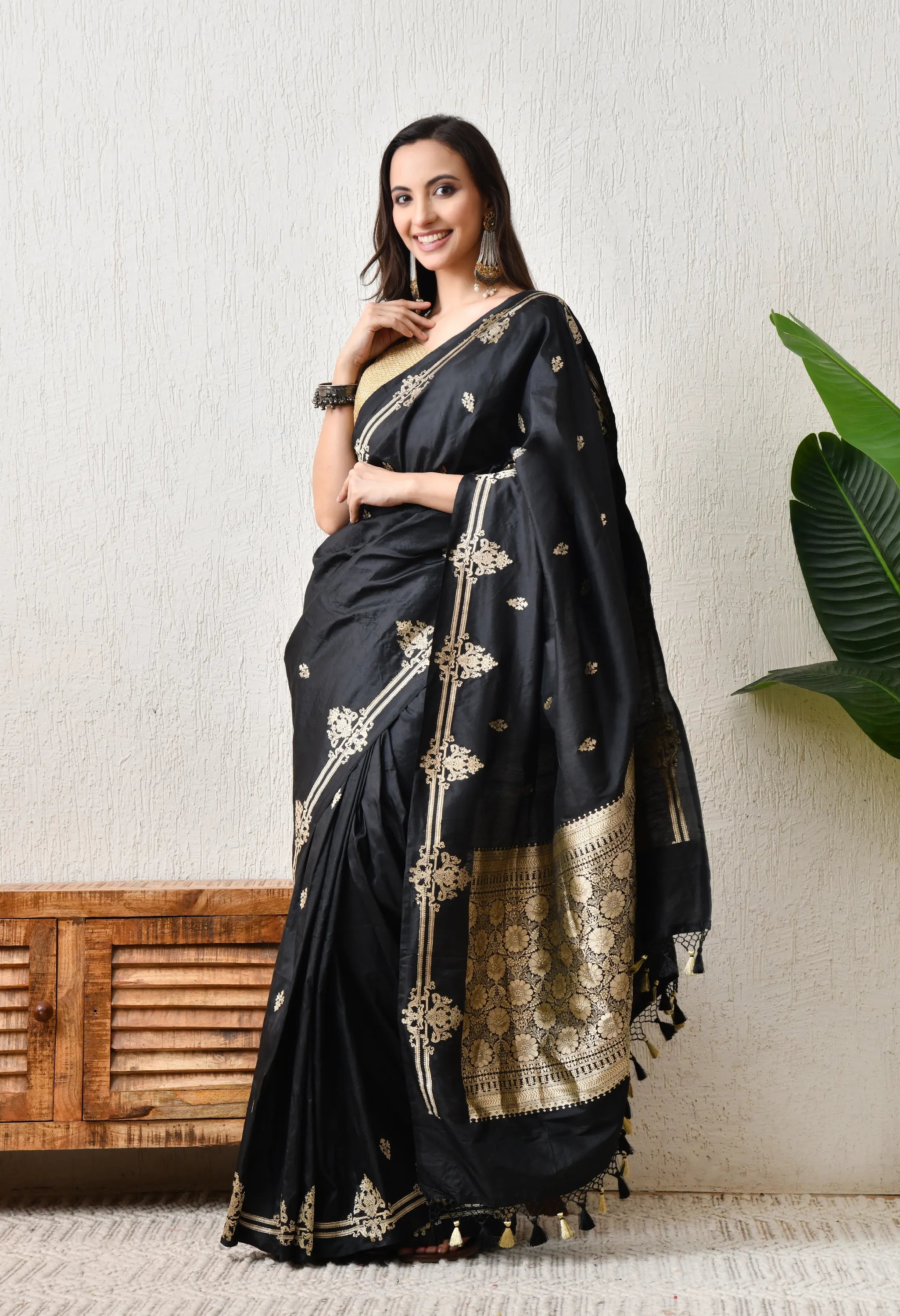 A graceful woman poses indoors wearing a pure Katan silk black saree from The Weavers Shop by Raavi, silk mark certified, with intricate gold zari work and delicate gold tassels on the pallu. The saree has elaborate vertical gold embroidery motifs on the front pleats and a rich, gold brocade pallu with floral and paisley patterns. The woman is smiling, standing beside a wooden cabinet, and is wearing matching gold jhumka earrings and bangles