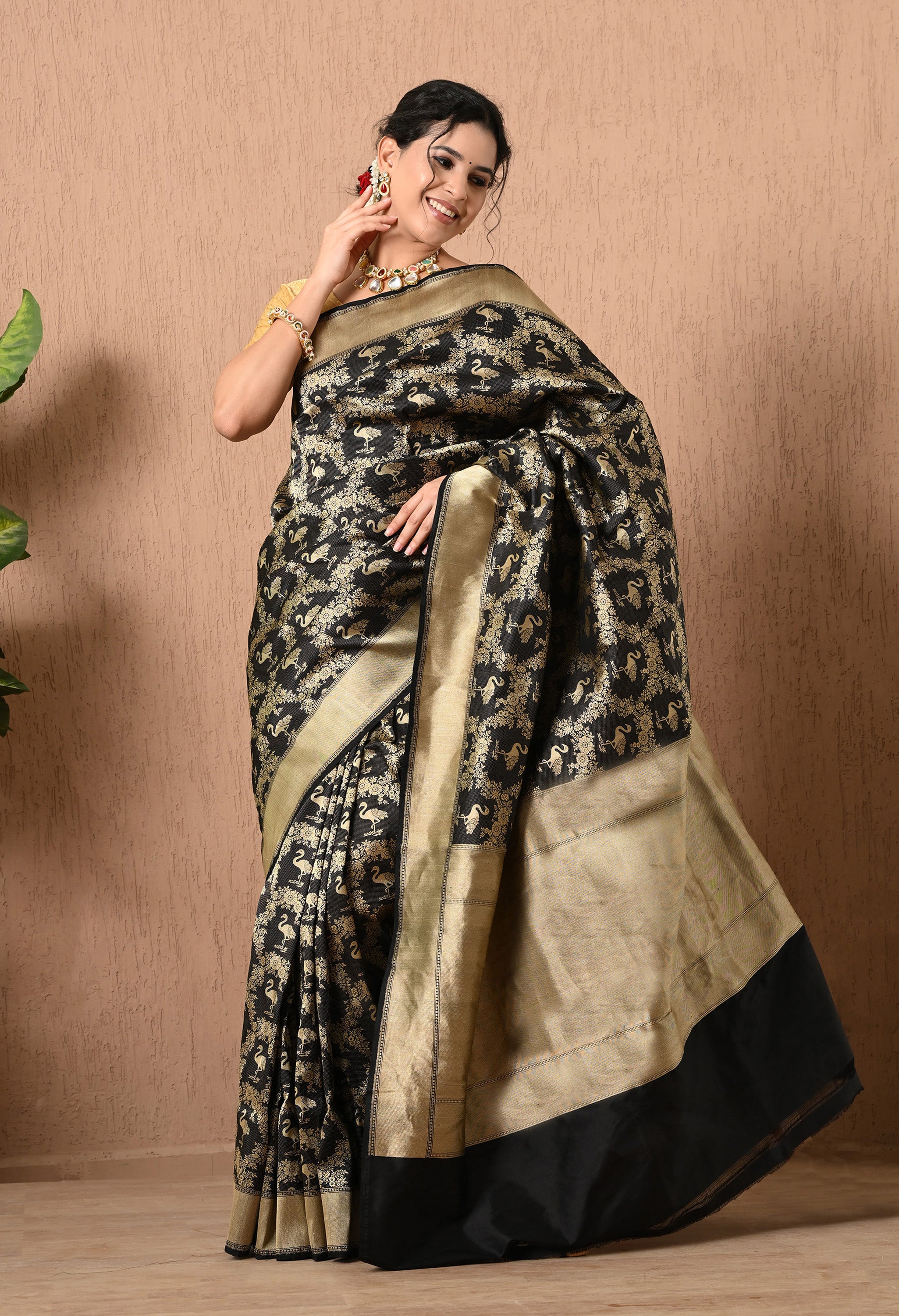 A full-length view of a woman smiling and posing in a black handwoven Shikarga saree on zari from The Weavers Shop by Raavi. The Pure Katan Silk fabric is adorned with a dense, shimmering gold pattern featuring traditional animal and floral motifs throughout. This Silk Mark Certified saree includes a wide, textured gold border and a solid black hem, paired with a gold blouse and a statement necklace.