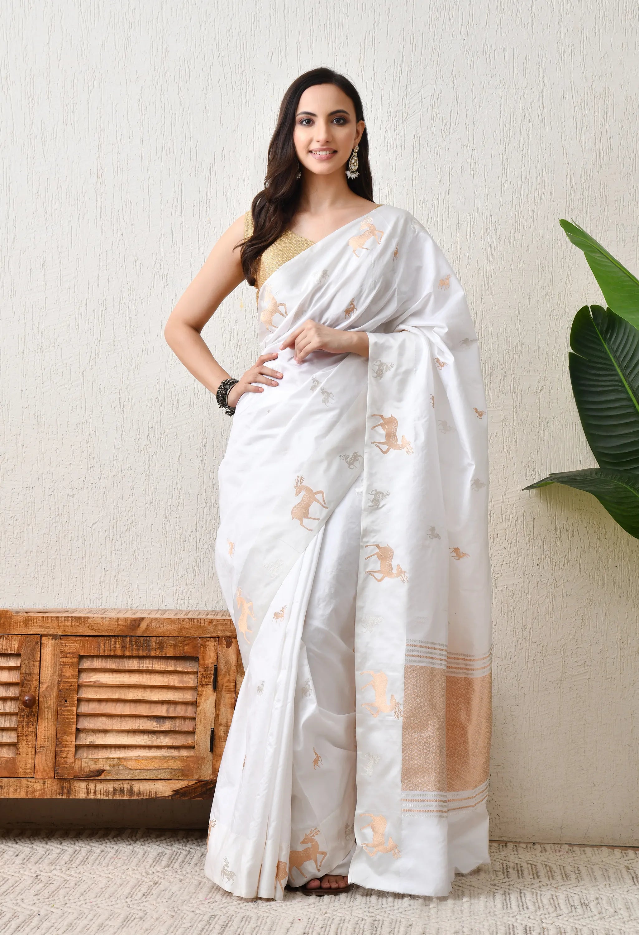A woman modeling a pristine white Hand Woven Pure Katan Silk saree from The Weavers Shop. The saree is adorned with elegant gold zari deer motifs scattered across the body and a coordinating zari border. This Silk Mark Certified garment is styled with a sleeveless gold blouse and traditional silver earrings, offering a sophisticated and royal look.