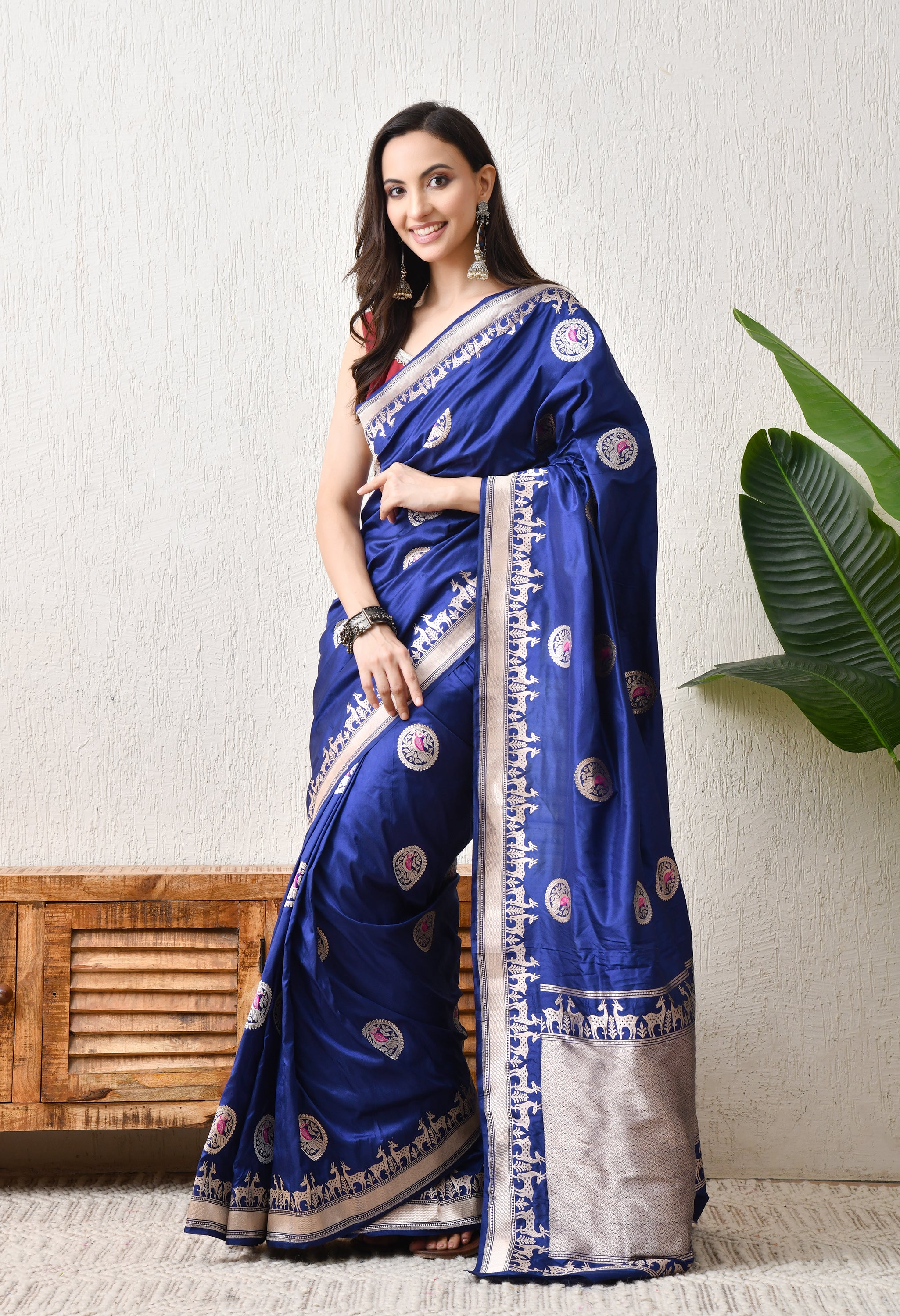 A side-frontal view of a woman posing in the royal blue Hand Woven Katan Silk saree from The Weavers Shop by Raavi. This angle highlights the rich luster of the Silk Mark Certified fabric and the repeating deer motifs of the Shikarga border. She is styled with traditional silver jewelry, showcasing the saree's suitability for royal festive or wedding wear.