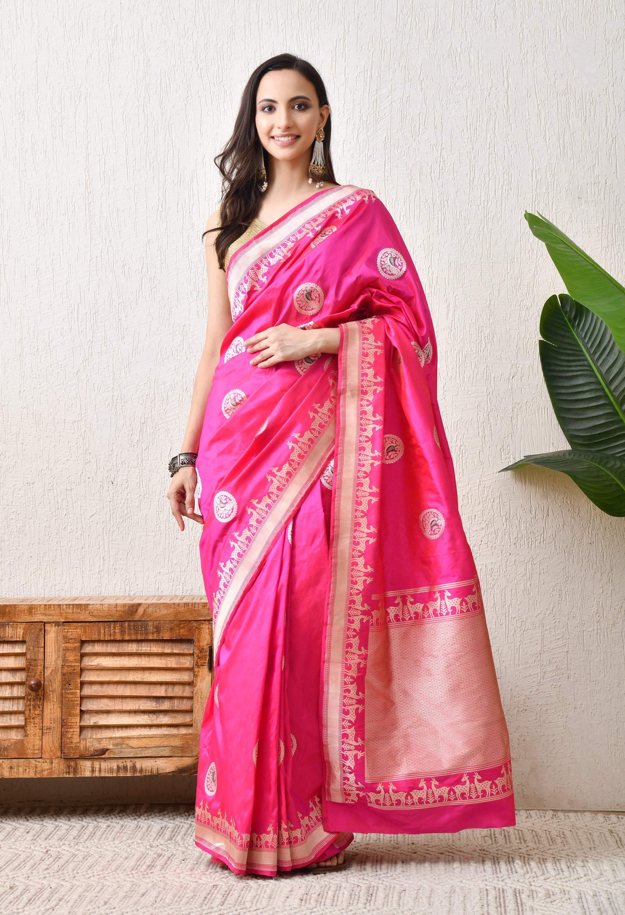 A woman modeling a vibrant pink Hand Woven Katan Silk saree from The Weavers Shop by Raavi. The saree features a traditional Shikarga border depicting intricate deer motifs and circular butis containing bird designs woven across the body. This Silk Mark Certified saree has a luxurious sheen and is paired with a gold blouse and statement earrings.