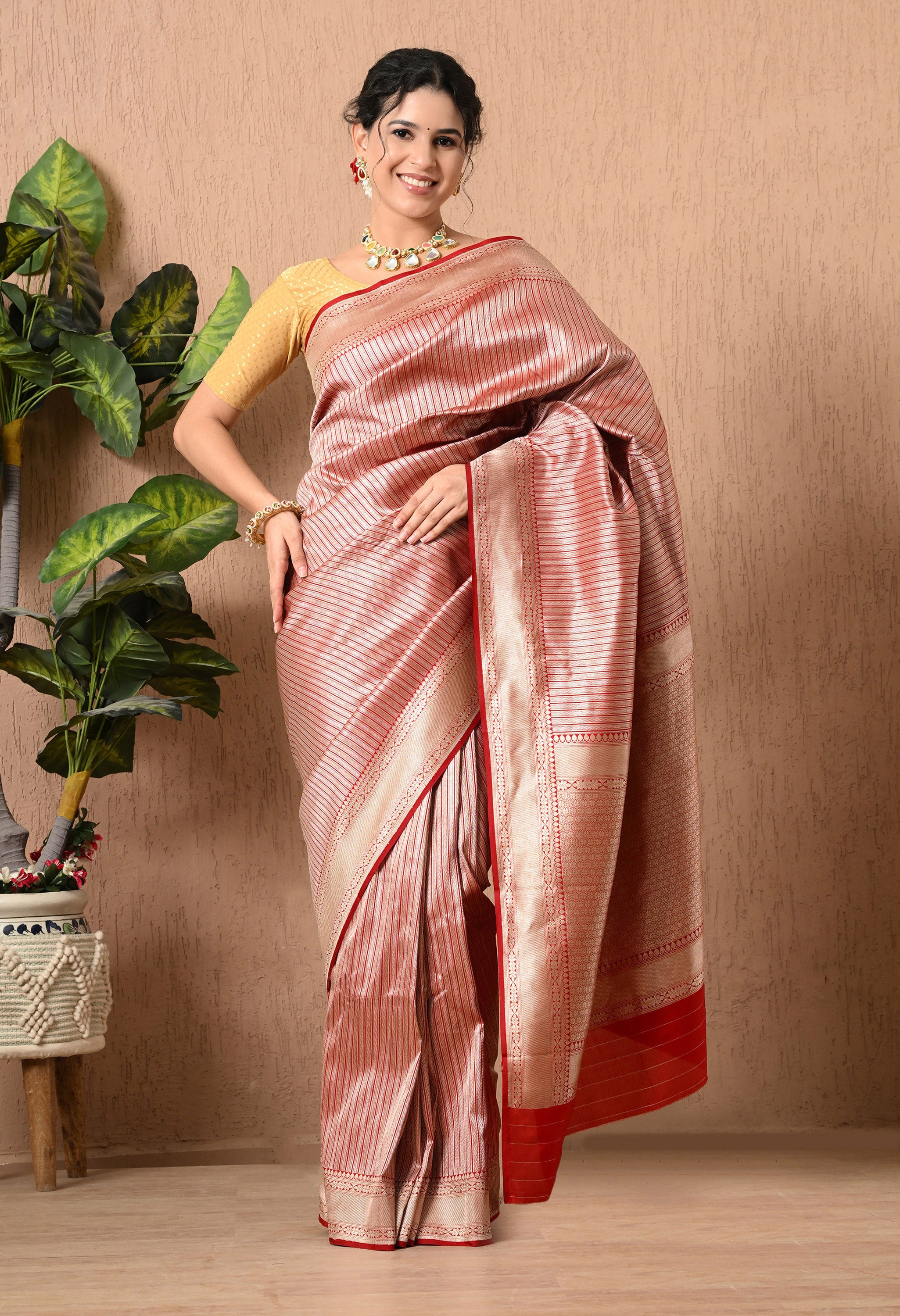 A full-length front view of a smiling woman posing in an elegant, Silk Mark Certified saree from The Weavers Shop by Raavi. The saree is crafted from premium Pure Katan Silk in a soft metallic or muted red hue. It features sophisticated hand-woven zari stripes that run vertically across the body of the garment. This authentic Banarasi piece is framed by a rich, solid red border at the hem and a broad, shimmering gold zari border and pallu. The look is completed with a gold textured blouse and a necklace