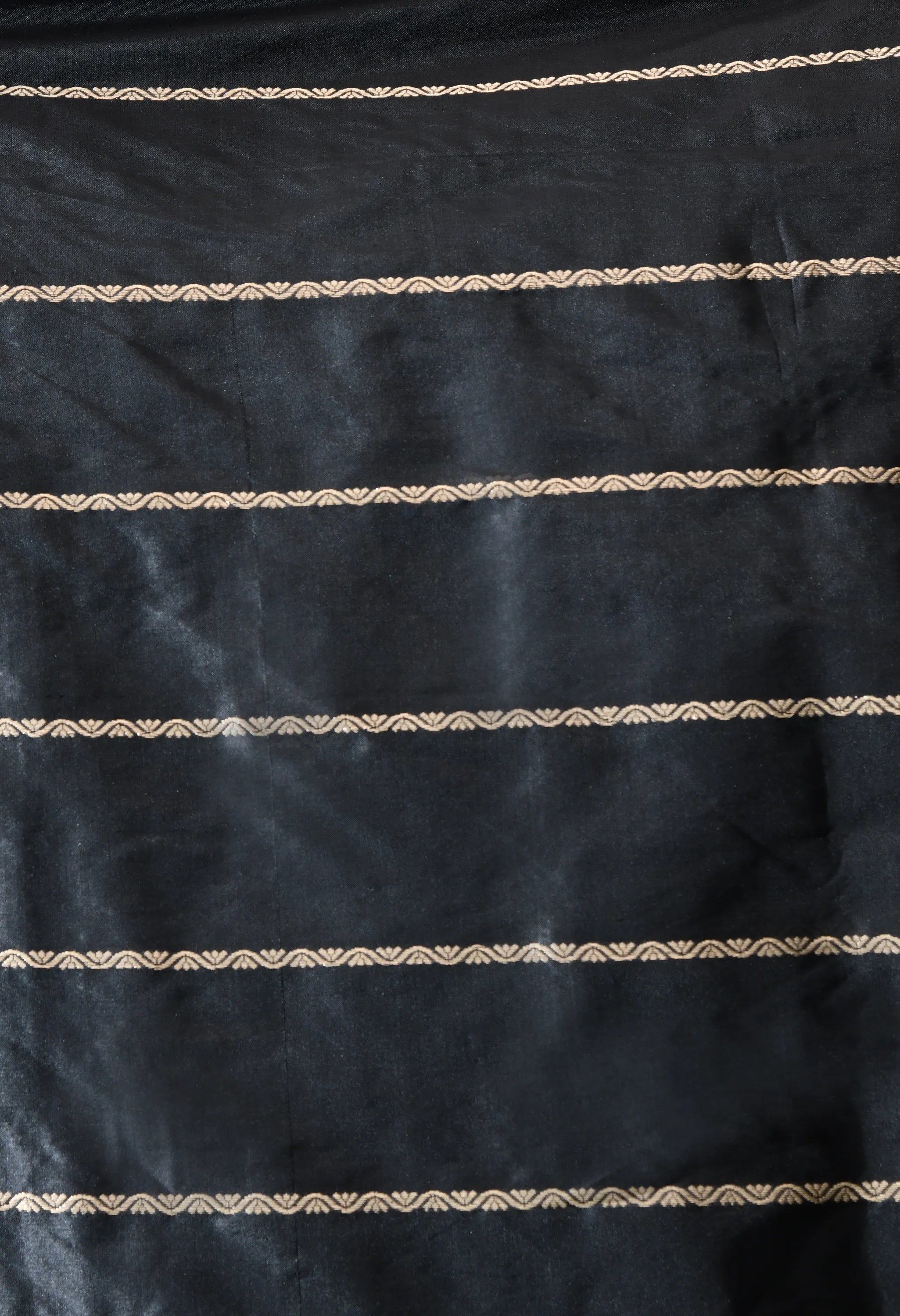 A close-up vertical shot of a Silk Mark Certified pure black silk blouse piece from The Weavers Shop by Raavi. The fabric features multiple horizontal rows of delicate, woven gold zari work in a continuous two  scallop pattern. The weave is tight and glossy, showcasing the subtle sheen of the black silk between the metallic gold horizontal lines.