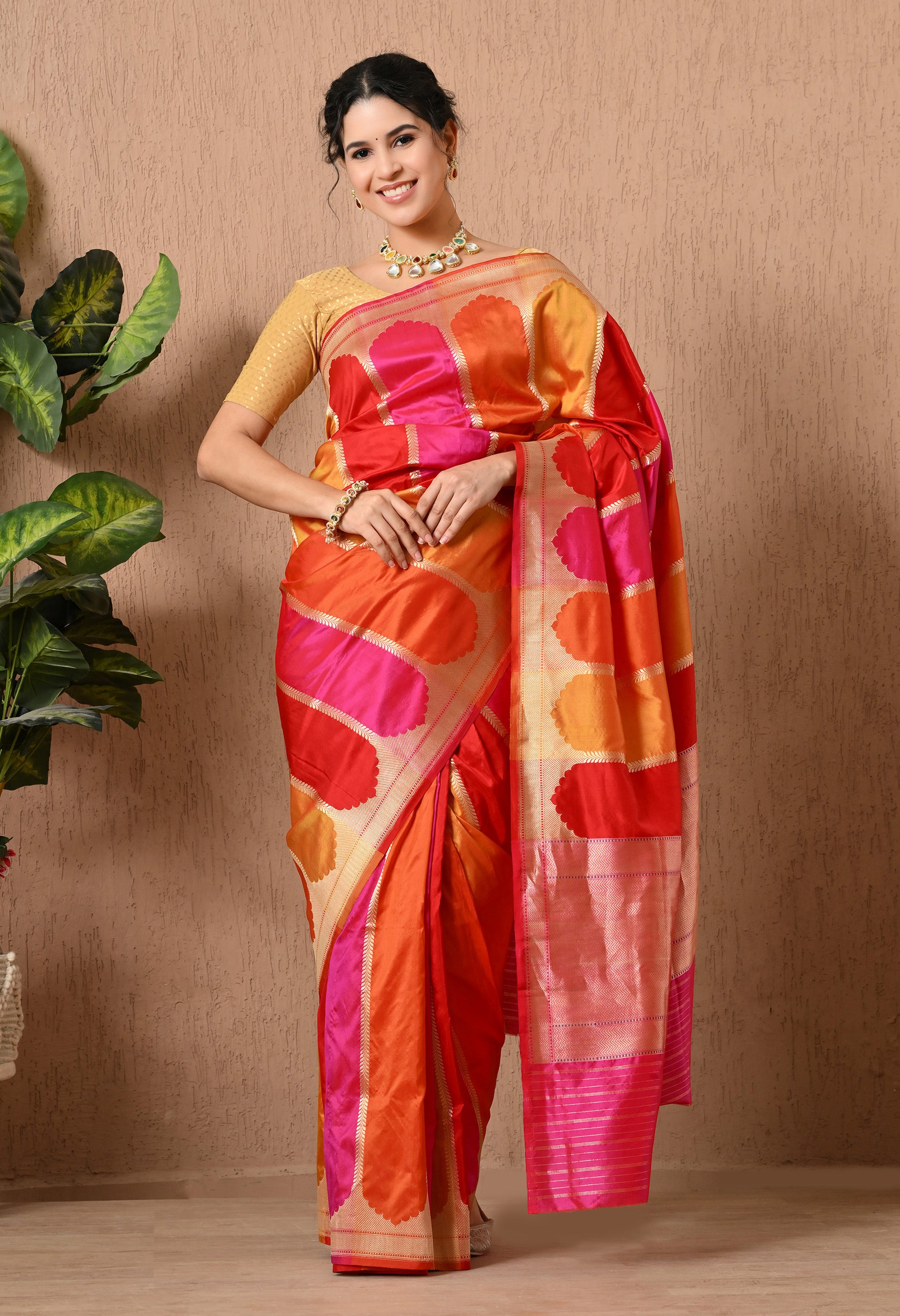 A woman stands facing forward, smiling and modeling a vibrant handwoven Pure Katan Silk saree from The Weavers Shop by Raavi. This authentic Banarasi garment showcases the traditional Rangkat weaving technique, featuring bold diagonal blocks of color in pink, orange, and red. The saree is adorned with intricate gold Zari work, including a detailed border and shimmering accents. This Silk Mark Certified piece is styled with a gold blouse and a statement necklace.
