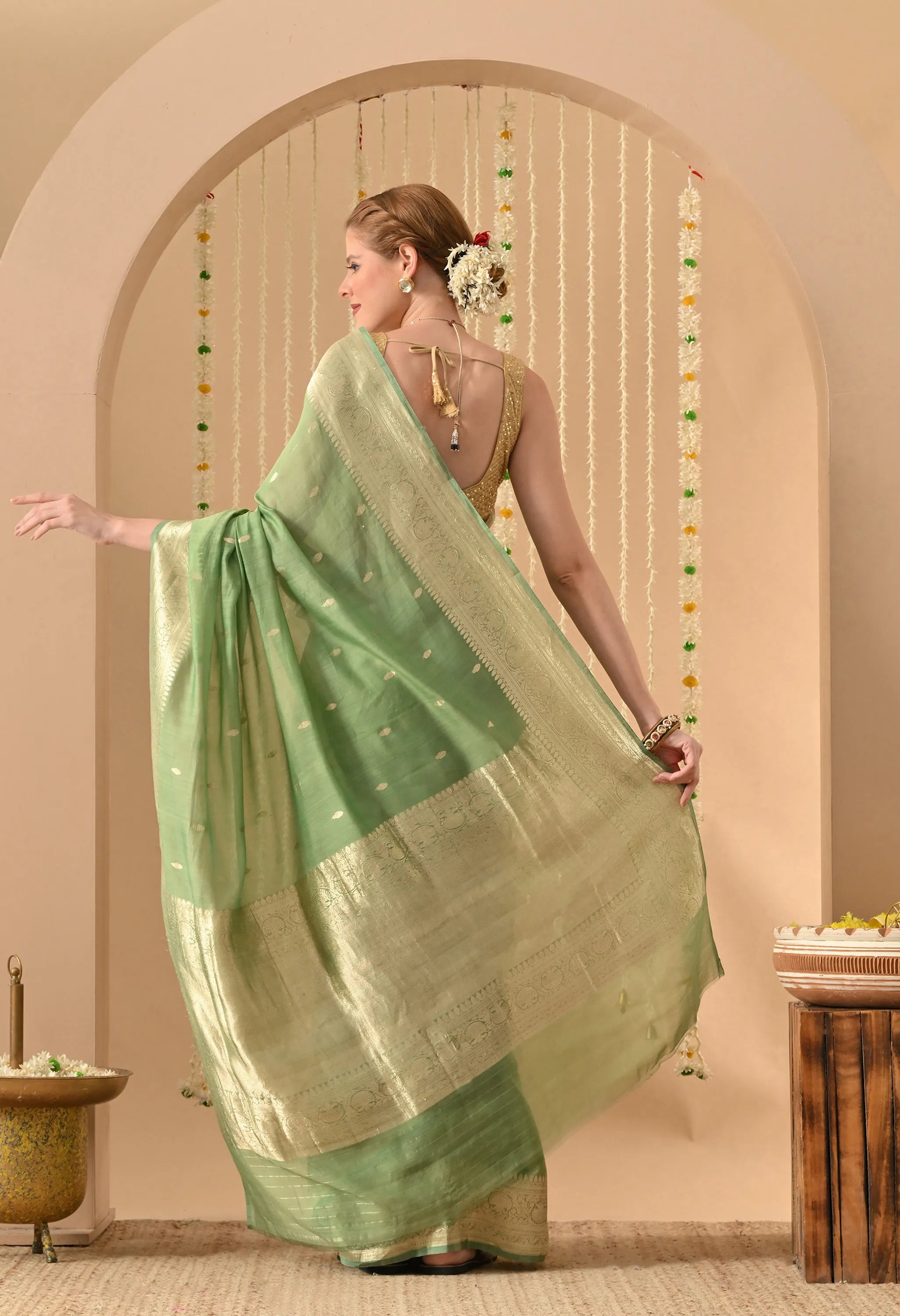 A rear-angle view of a woman showcasing the elegant drape and detailed pallu of a Light Green Pure Silk Banarasi Saree from The Weavers Shop by Raavi. This hand-woven garment features the labor-intensive Karuwa Weave, with delicate gold zari work butis (motifs) appearing across the high-quality silk. The broad, shimmering gold zari border and pallu are prominently displayed as the model extends the fabric, revealing a refined floral pattern. She is styled with a sleeveless gold blouse.