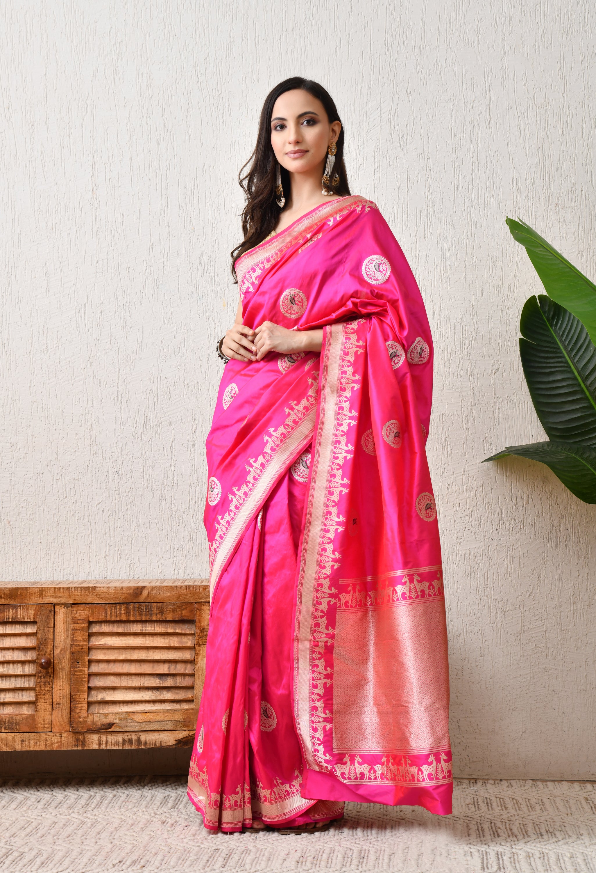 A side-frontal view of a woman posing in the pink Hand Woven Katan Silk saree from The Weavers Shop by Raavi. This angle highlights the rich luster of the Silk Mark Certified fabric and the repeating deer motifs of the Shikarga border. She is styled with traditional jewelry, showcasing the saree's suitability for wedding or royal festive wear.