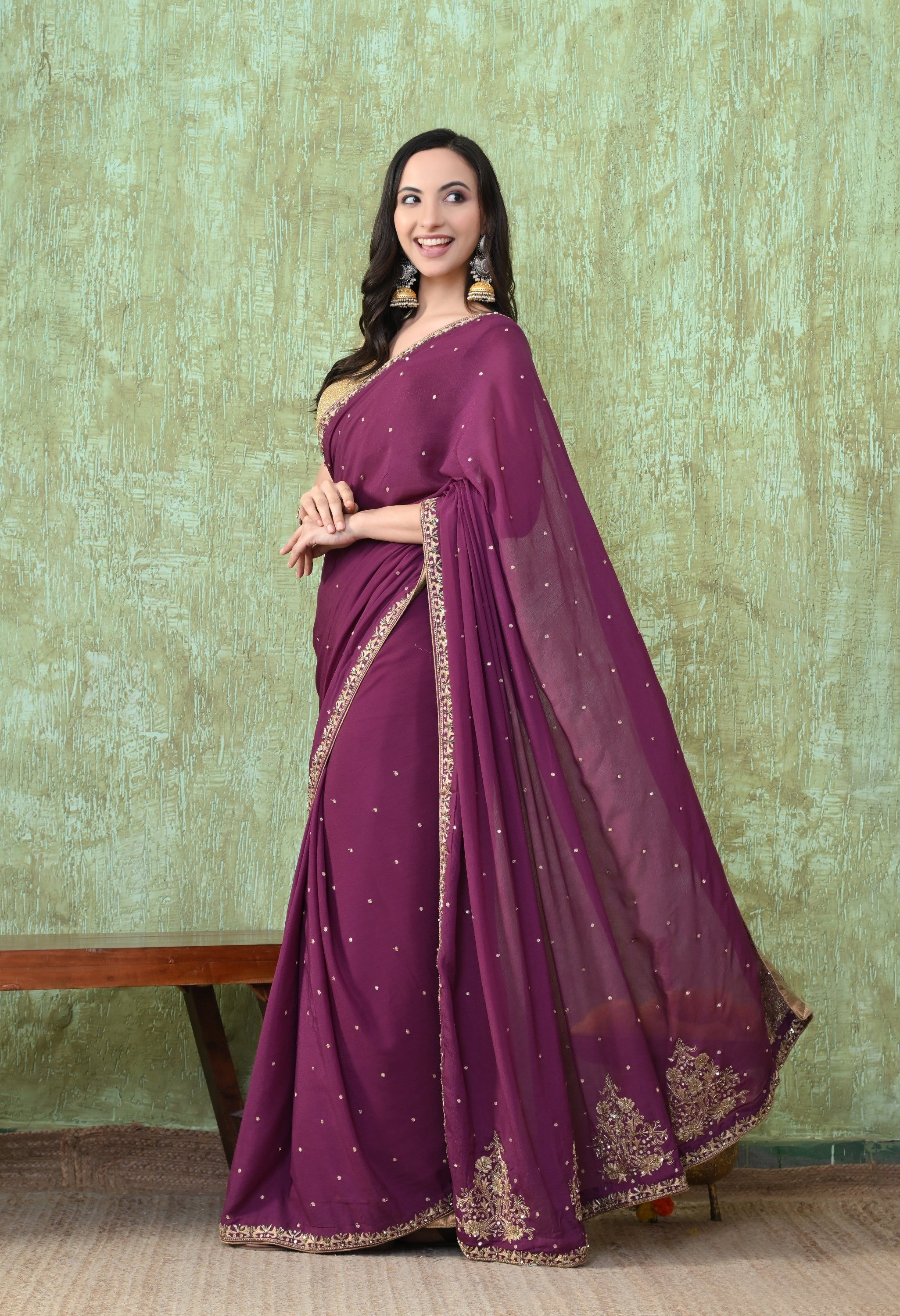 A side-angle view of a woman posing in a rich wine-colored crepe saree featuring detailed zardozi handwork by The Weavers Shop by Raavi. The gold floral embroidery is prominently displayed along the base and borders of the saree. She is smiling and wearing a gold blouse and statement earrings, standing against a textured pale green wall.