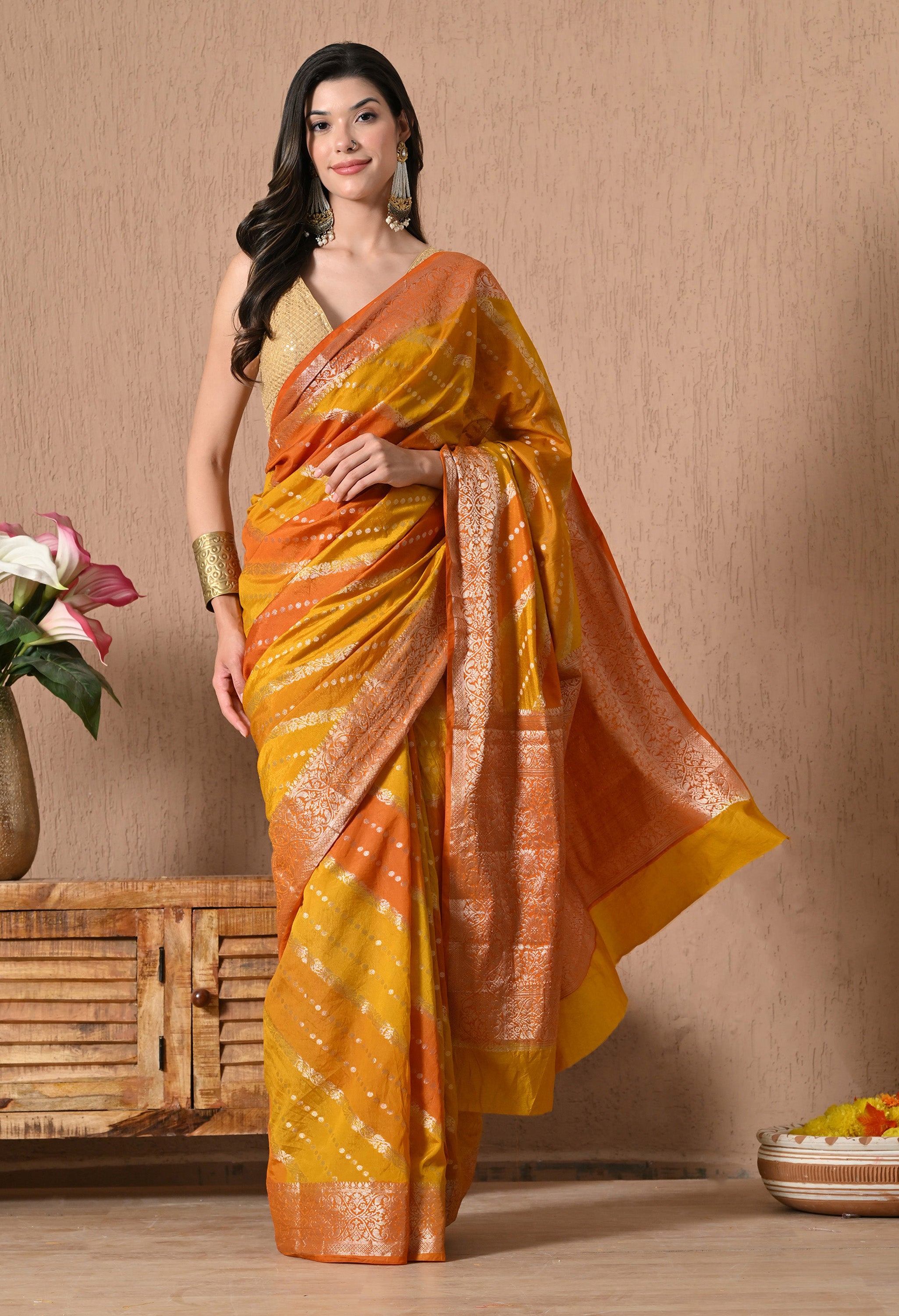 A model poses in a vibrant Rangkat Banarasi silk saree from The Weavers Shop, showcasing a sunset-inspired palette of mustard yellow and deep orange diagonal stripes. The saree features intricate golden zari work with traditional geometric and traditional motifs on the borders and pallu, accented by small gold-toned woven dots across the body. She is styled with a sleeveless gold blouse and traditional earrings 