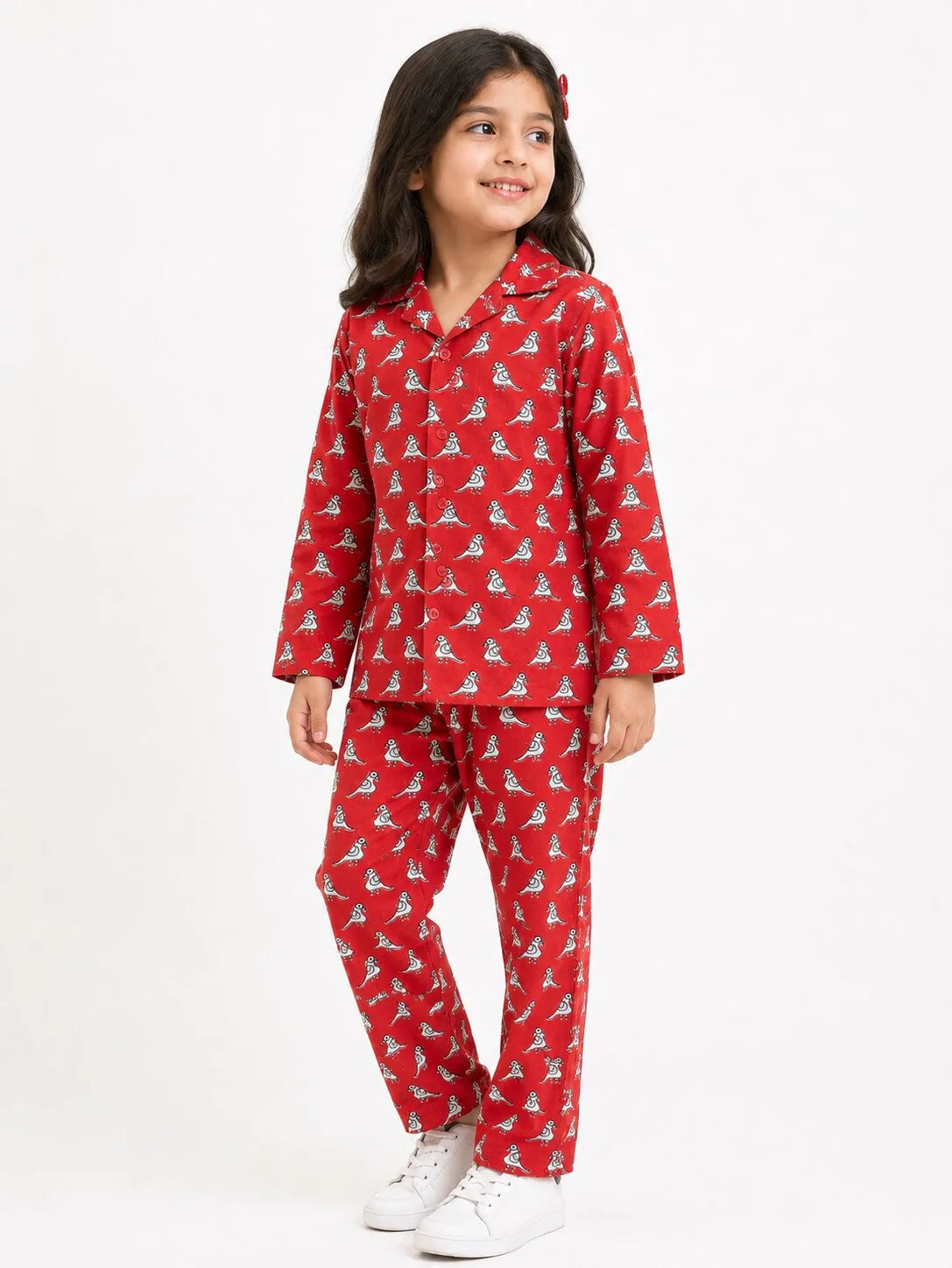 A young girl smiling while wearing a red long-sleeved pajama set featuring a white all-over bird motif, standing against a dark background