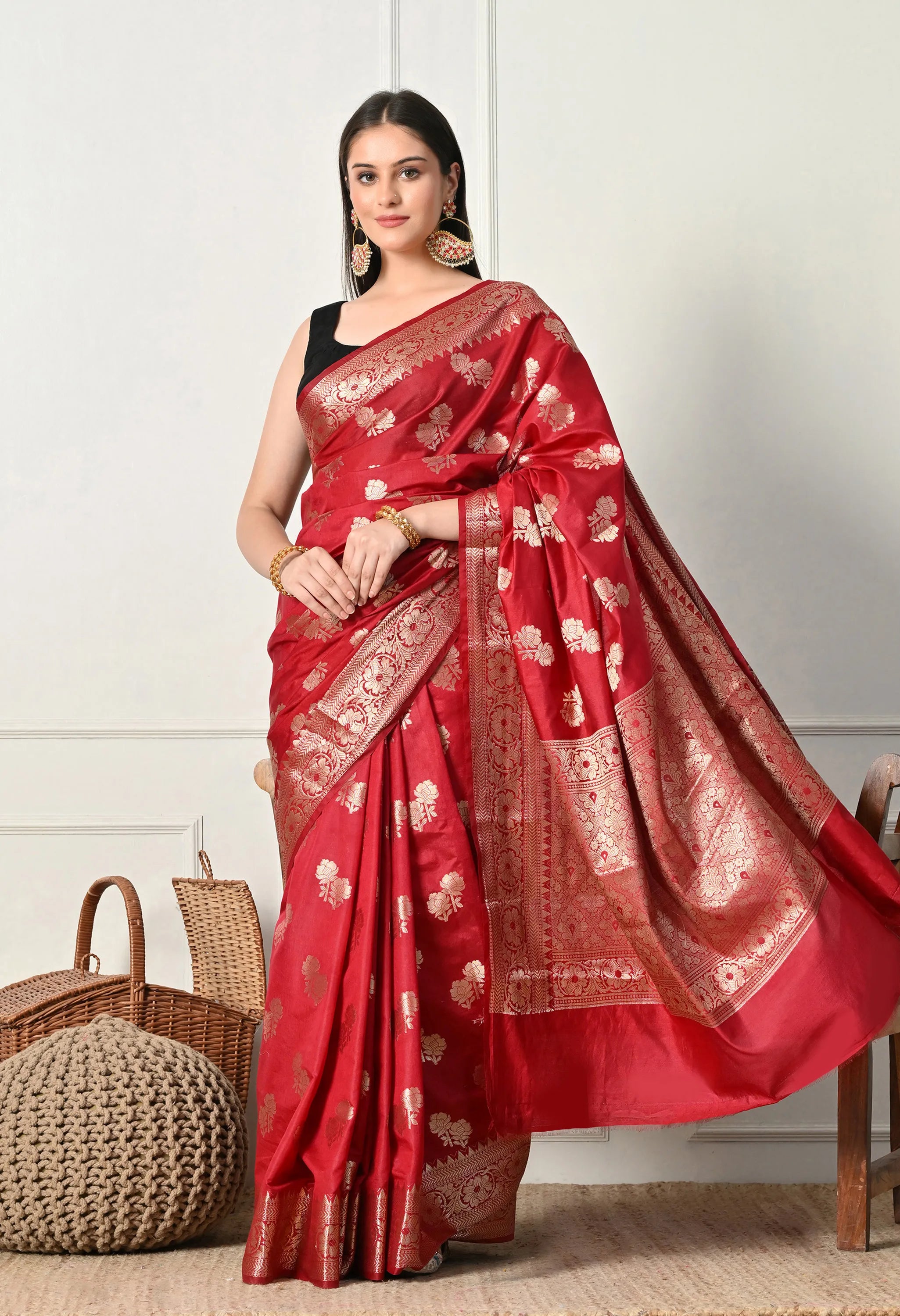 A full-length front view of a woman posing in a luxury red silk Banarasi saree featuring heavy golden zari work. The saree is draped to show the decorative pallu with detailed  floral patterns. She is styled with a black sleeveless blouse and gold jewelry, standing in a bright room with minimalist decor, including a wicker basket and a woven pouffe in the background.