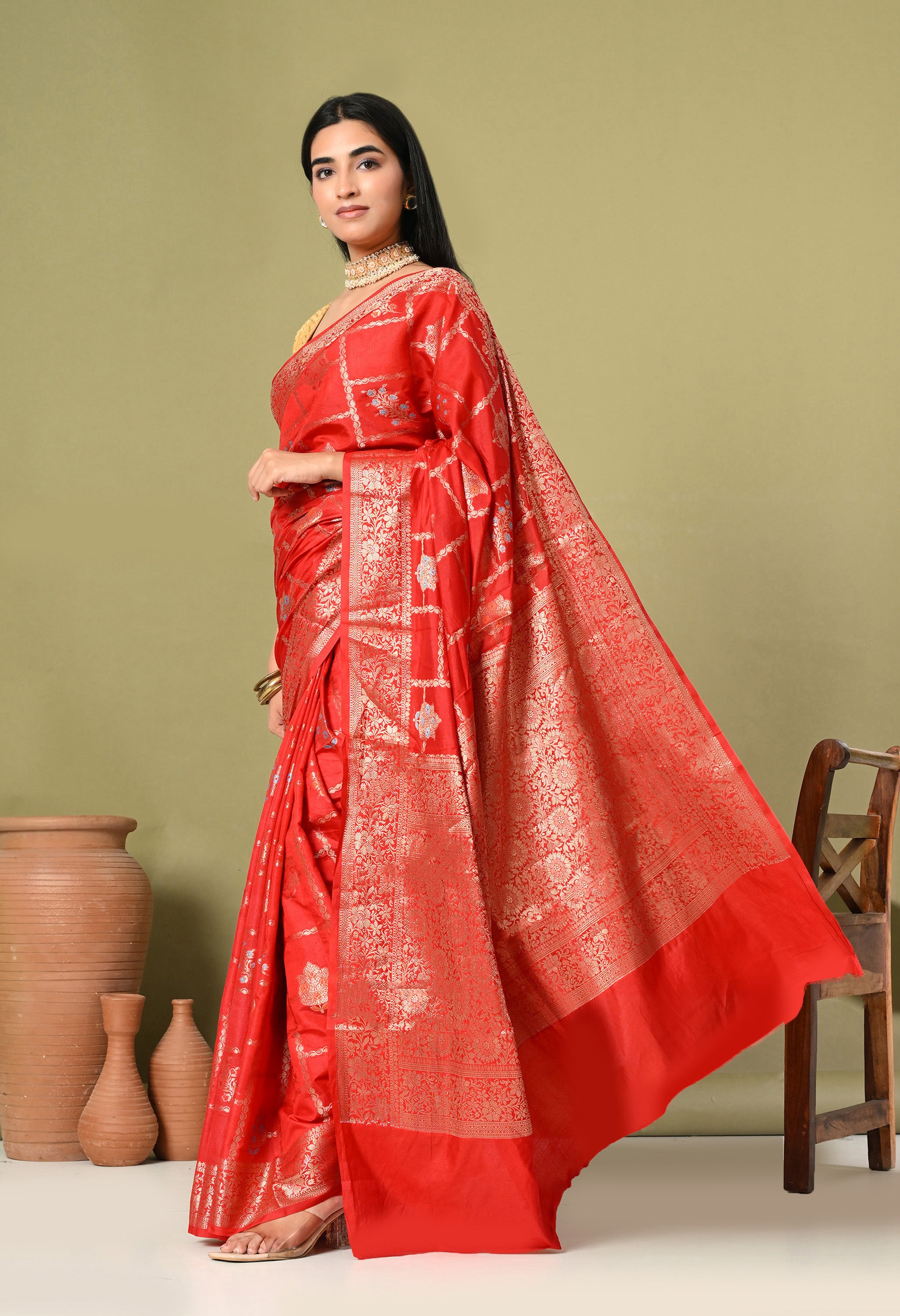 A side profile of a woman showcasing the fluid drape and luxurious texture of a The Weavers Shop by Raavi Banarasi saree. This angle emphasizes the wide gold zari border and the repeating Tilfi embroidery across the red silk. The styling, featuring a gold-textured blouse and traditional accessories, reflects the sophisticated wedding and event wear offered by The Weavers Shop by Raavi.