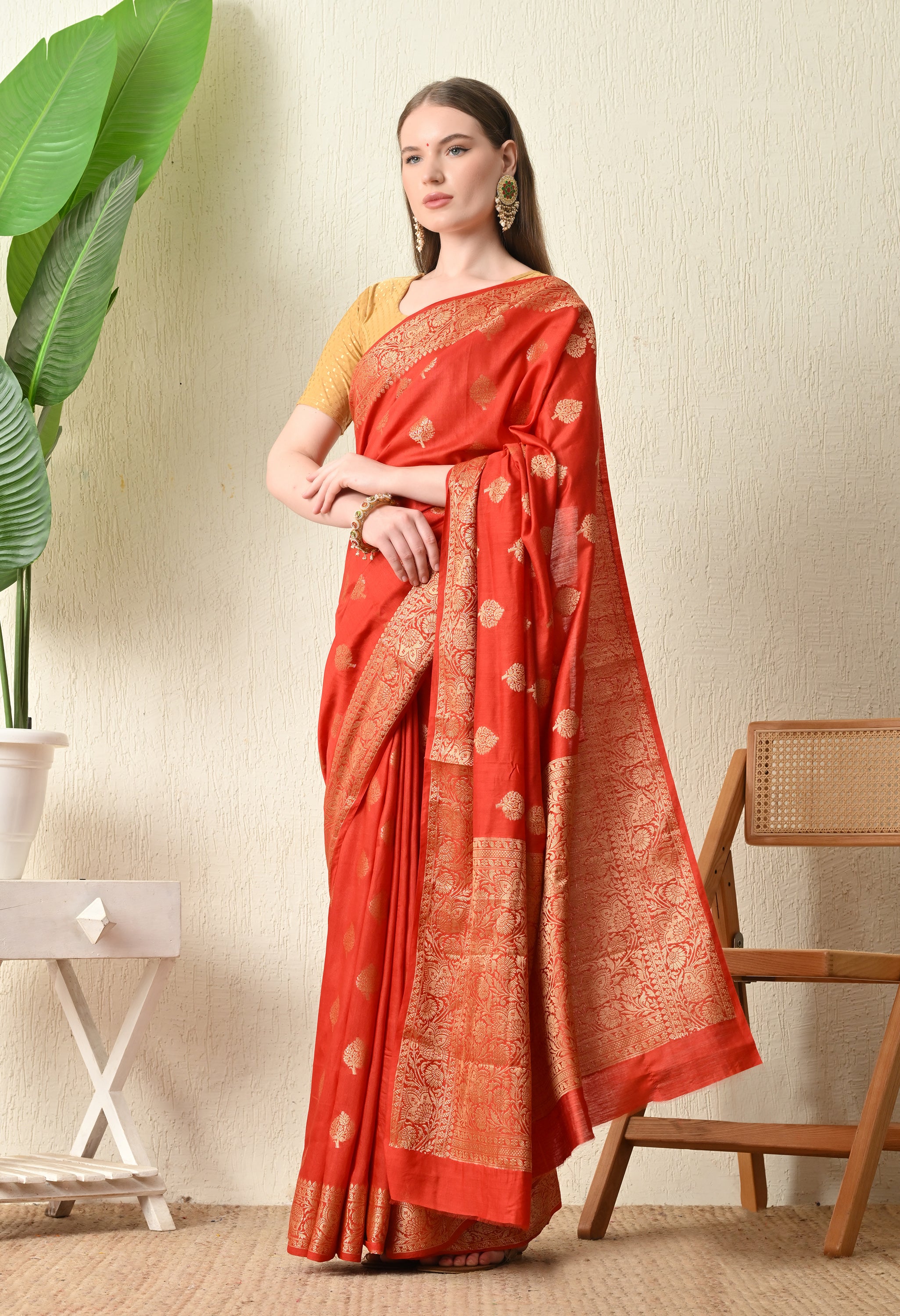 woman poses in a vibrant red Banarasi silk saree from The Weavers Shop by Raavi. The saree features intricate gold zari work, including traditional floral leaf motifs (butis) scattered across the body and a wide, ornate gold border. She is wearing a contrasting short-sleeved gold blouse and traditional Indian jewelry, standing against a neutral textured wall.