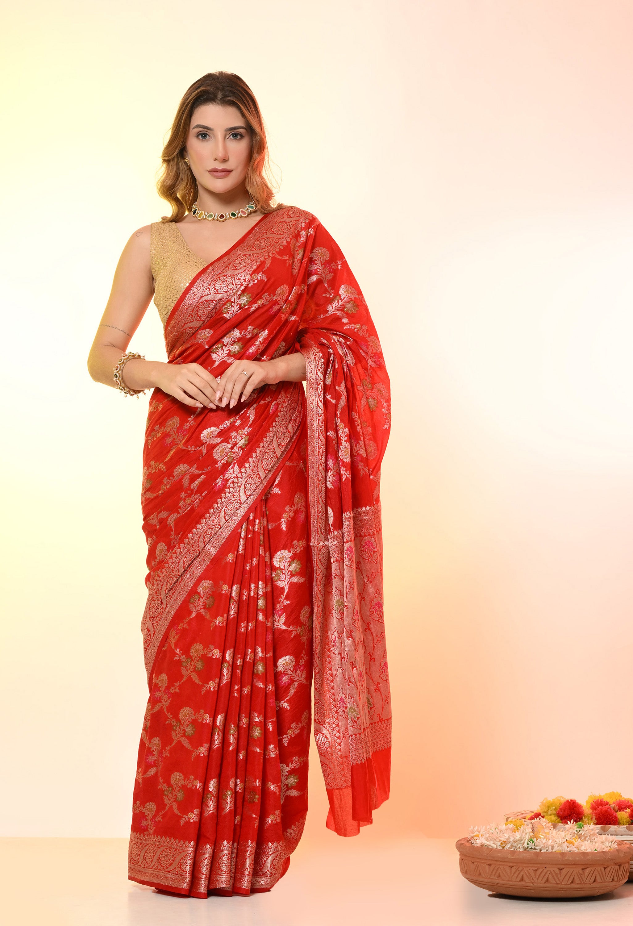 A woman poses in a vibrant red Banarasi-style silk-blend saree from The Weavers Shop by Raavi, featuring intricate Tilfi work and shimmering silver zari floral patterns. This elegant machine-woven drape is styled with a sleeveless gold blouse and a delicate choker necklace, set against a soft gradient background with a traditional decorative floral bowl.