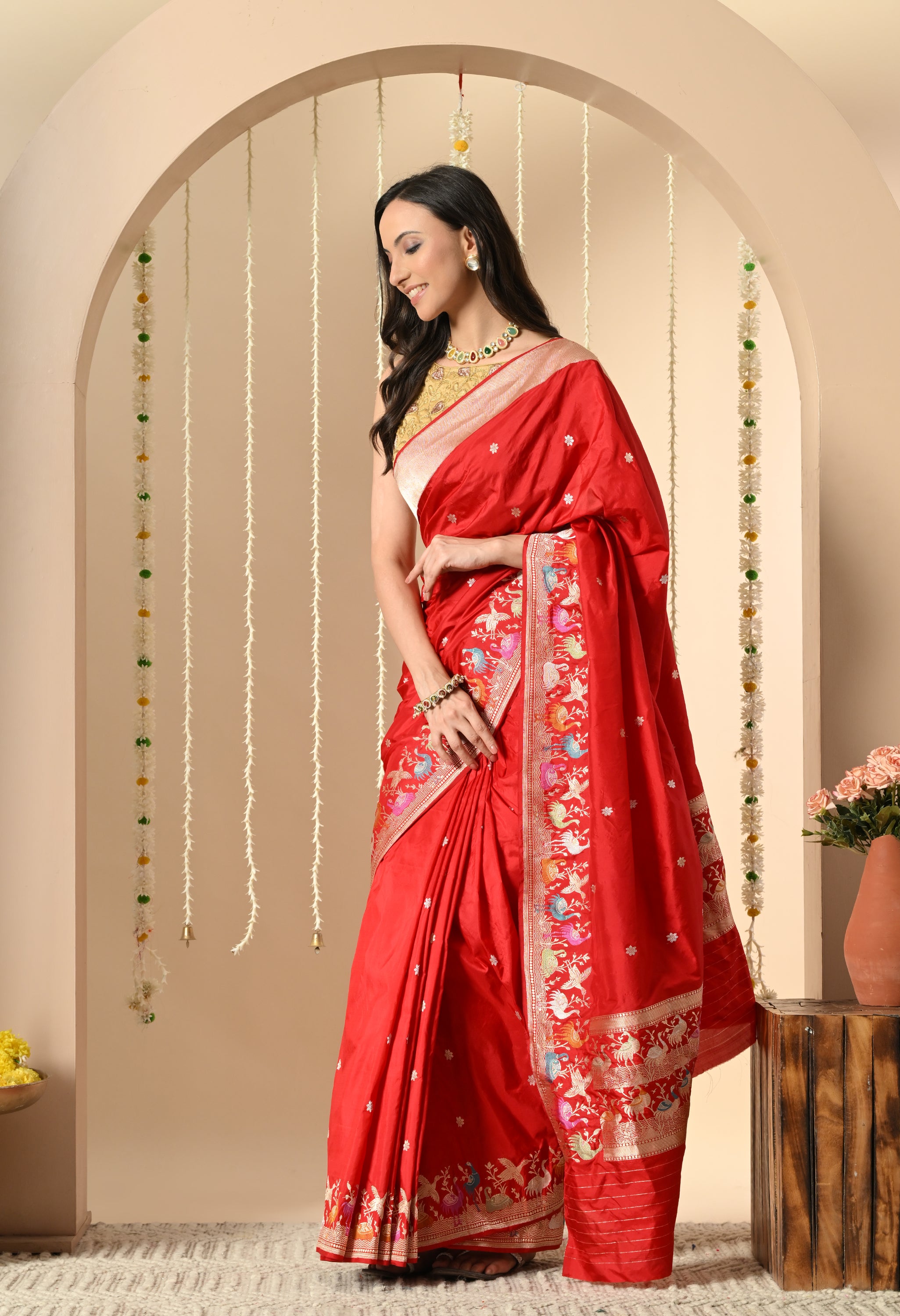 A beautiful woman is modeling a vibrant red Katan silk saree from The Weavers Shop by Raavi in an arched setting. The Silk Mark certified garment features a detailed, multicolored Shikarga border with woven motifs of animals and figures, and is paired with a gold blouse.