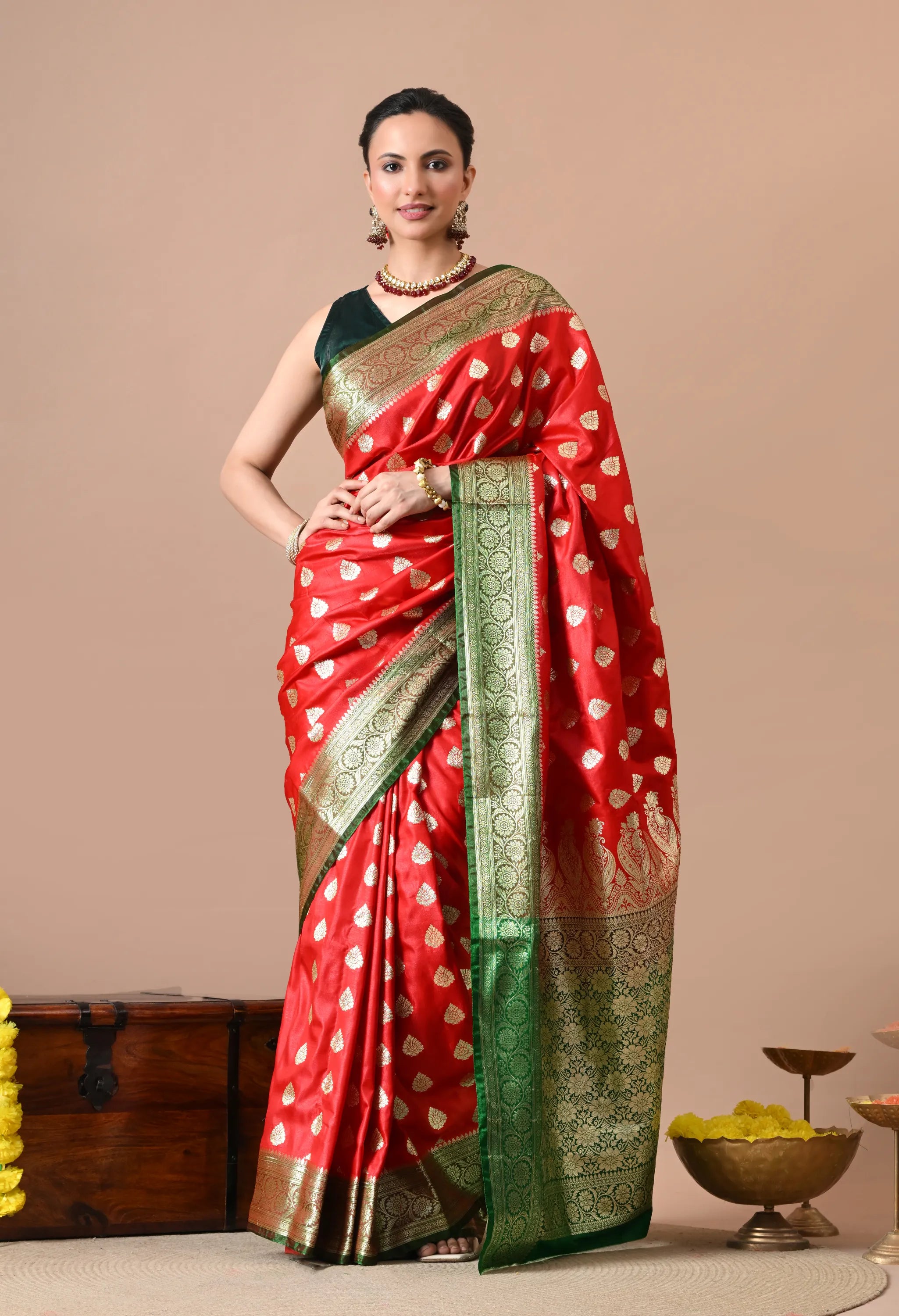 A woman posing in a vibrant red silk-blend saree with gold-toned leaf motifs and a wide green zari border, paired with a dark green sleeveless blouse.