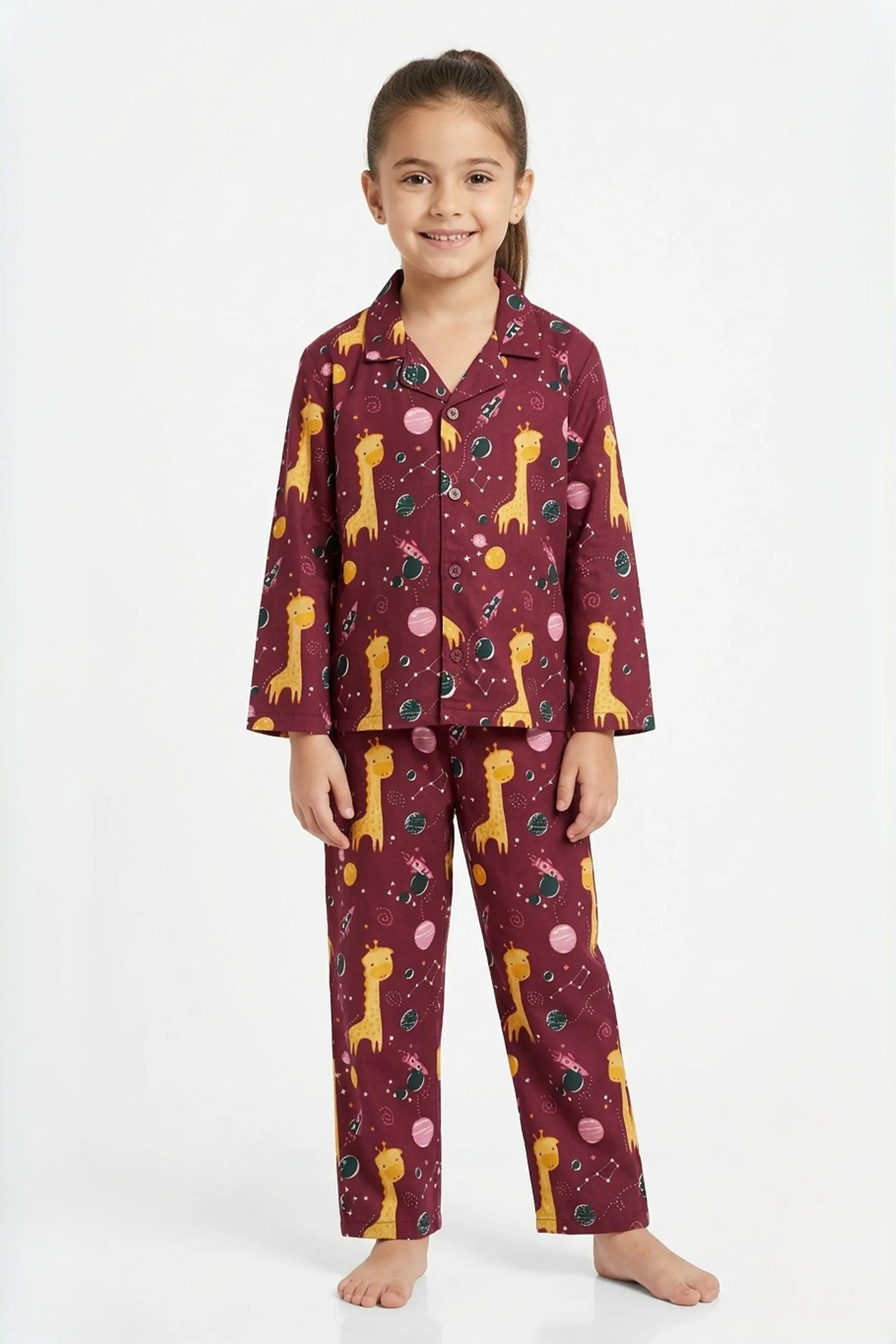 A young girl standing against a white background, wearing a maroon long-sleeved button-up pajama set with a yellow giraffe animal print