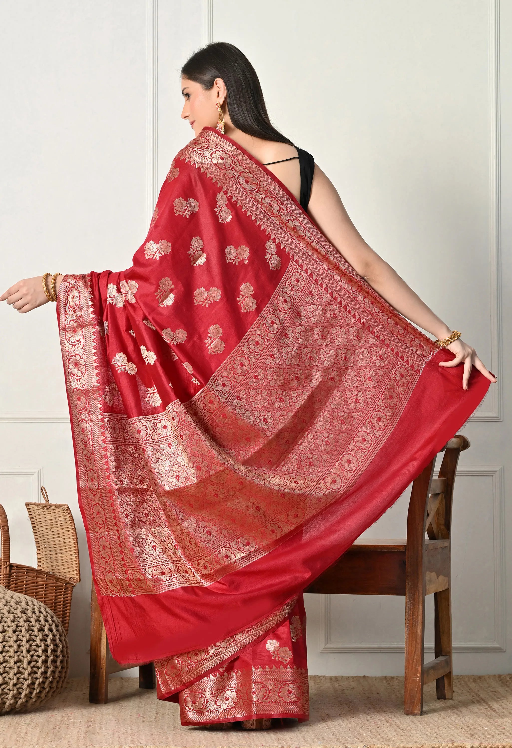 A back-angle view of a woman extending the heavy golden zari pallu of a red silk Banarasi saree to display its full, intricate design. The wide border and dense floral patterns in the gold thread are prominent against the rich red silk fabric. The shot also shows the back of her black sleeveless blouse and the elegant flow of the saree’s drape.