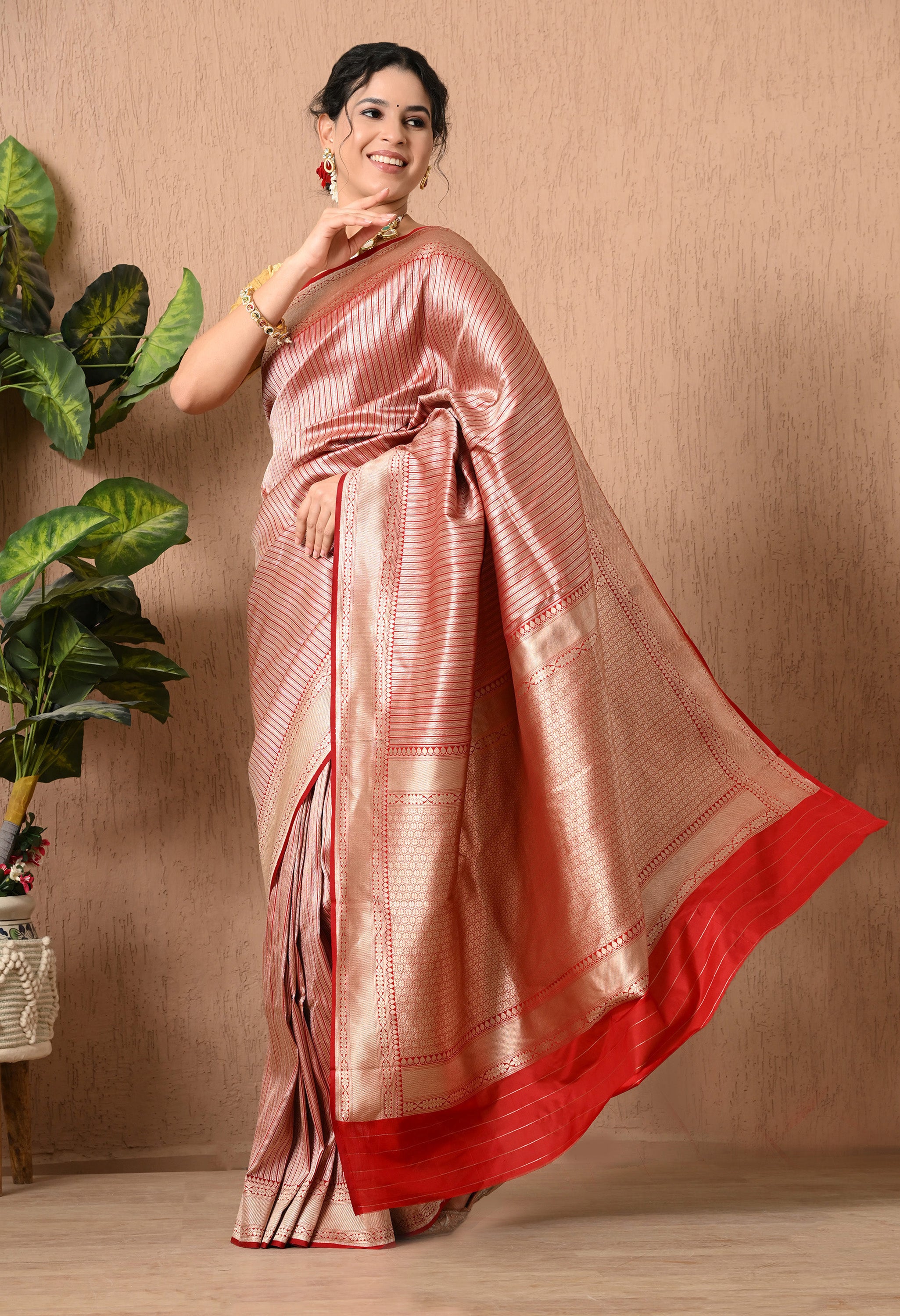 A full-length side-profile view of a woman posing in a Silk Mark Certified saree from The Weavers Shop by Raavi. The saree is made of premium Pure Katan Silk in a metallic red and silver tone, featuring hand-woven vertical zari stripes. The image captures a dynamic "flying pallu" effect, where the broad, intricate gold zari end-piece of the saree is billowing gracefully behind her, showcasing the detailed craftsmanship and fluid drape of the fabric. She is styled with a gold blouse, jewellry 
