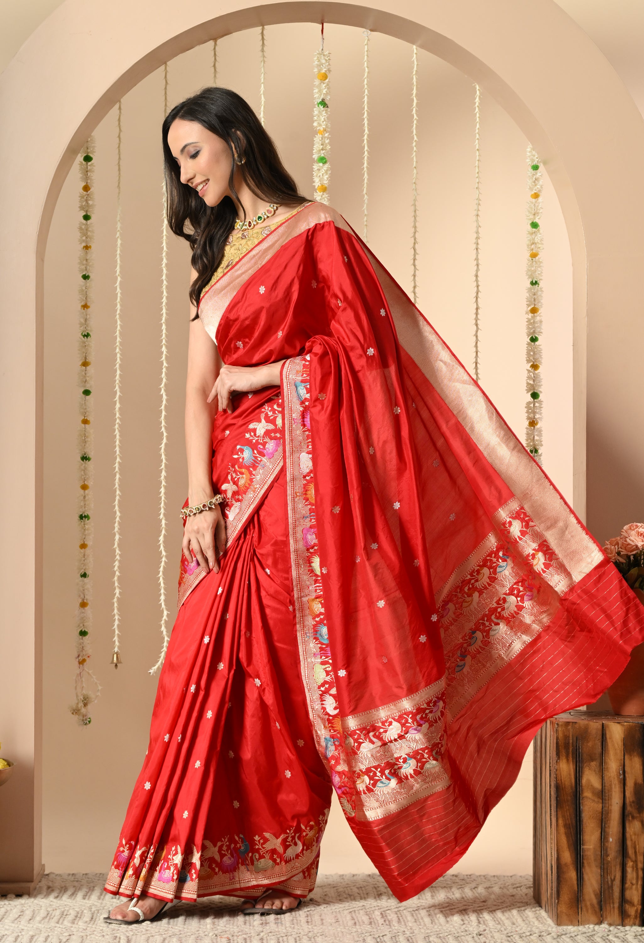 A woman is captured in a side profile pose, modeling a vibrant red Katan silk saree from The Weavers Shop by Raavi, which is Silk Mark certified. The saree features a wide, multicolored Shikarga border with intricate woven motifs of animals and figures, and the body of the saree has subtle Zari butis. The scene is set within a cream-colored arch.