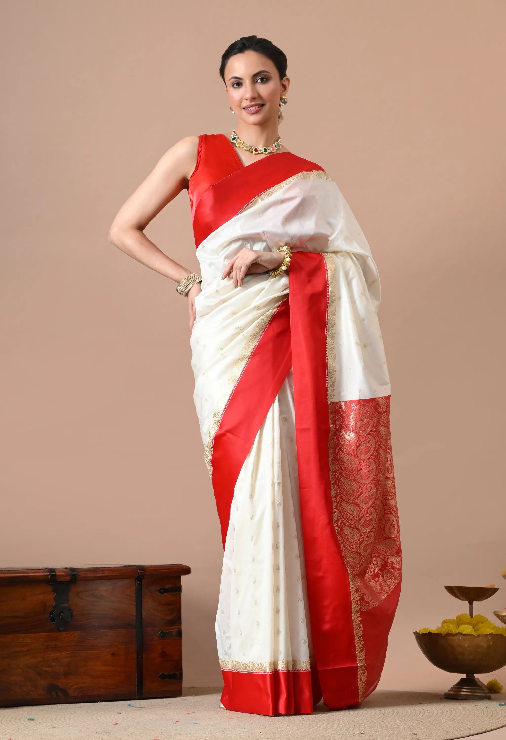 Woman posing in a white silk blend saree with red woven borders and pallu, paired with a red sleeveless blouse.
