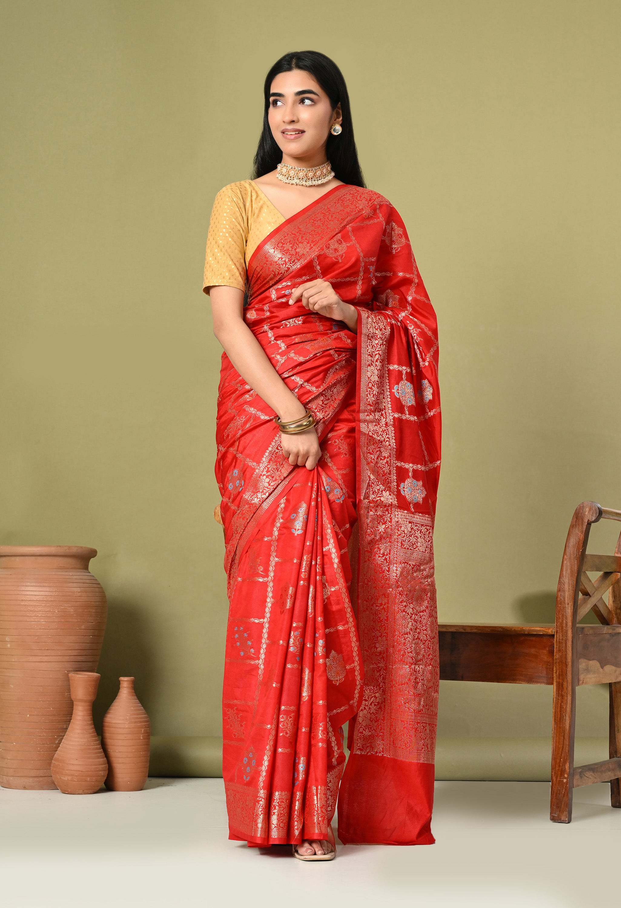 A full front view of a woman elegantly draped in a red Tilfi Banarasi silk saree from The Weavers Shop by Raavi. The saree features a meticulous gold grid design with alternating floral motifs, paired with a matching gold blouse and a pearl choker necklace. This shot highlights the complete ensemble and the vibrant, luxurious finish of the silk offered by The Weavers Shop by Raavi.