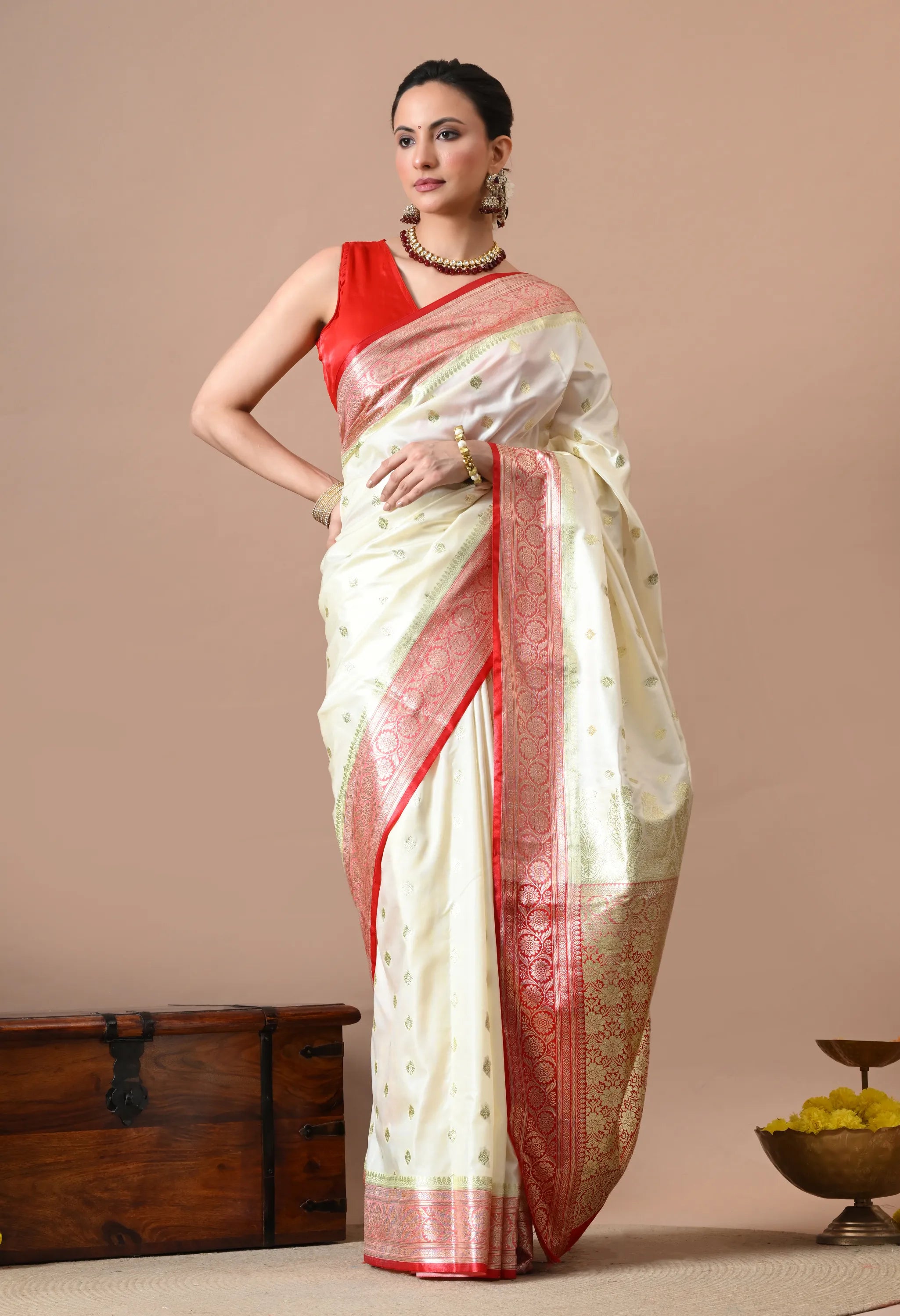 Woman posing in an off-white silk blend saree with a red and gold woven border and matching sleeveless red blouse.
