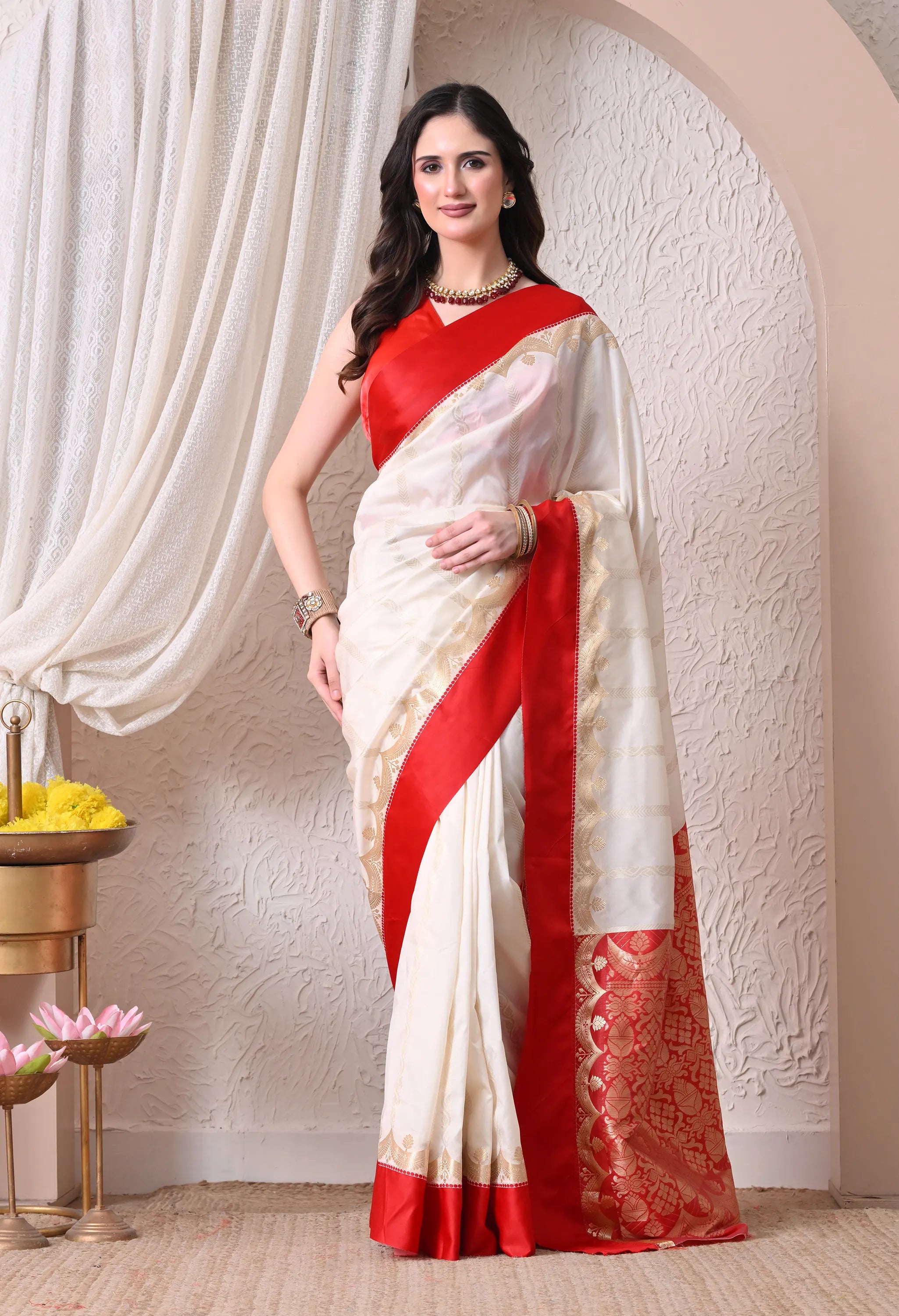 A woman modeling a traditional off-white silk blend saree accented with a striking crimson red border and a grand golden zari woven pallu.
