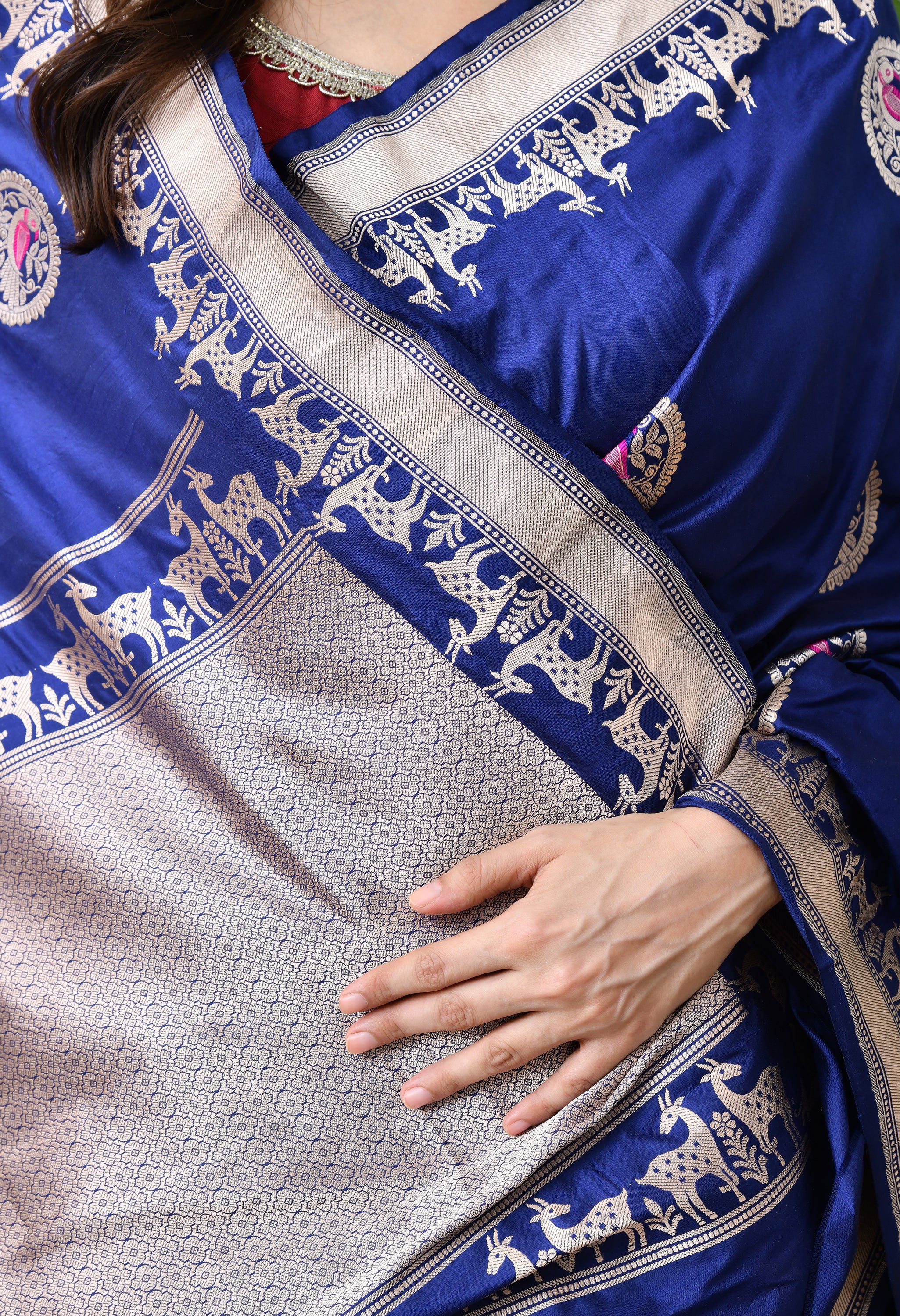 A detailed close-up shot of the torso and hands of a woman draped in a royal blue Hand Woven Katan Silk saree by The Weavers Shop by Raavi. The image provides a clear view of the intricate silver zari Shikarga border featuring rows of deer and floral patterns, alongside circular butis containing bird designs. This Silk Mark Certified saree is paired with a maroon blouse featuring a silver embroidered neckline.