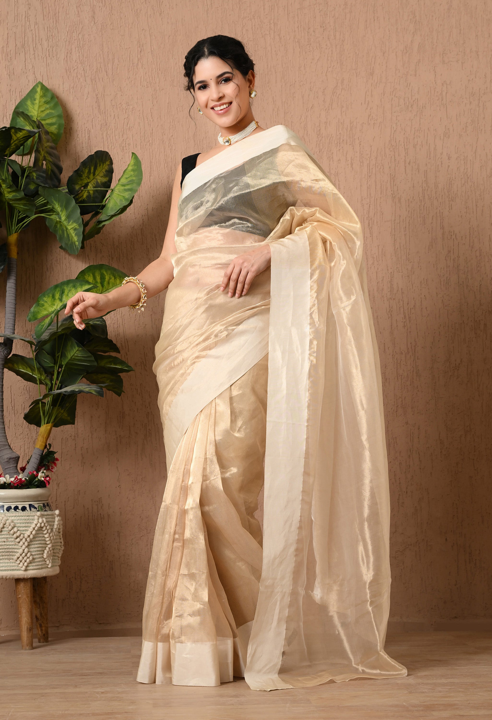A side-angle view of a woman posing in a light golden handwoven tissue saree from The Weavers Shop by Raavi. The image highlights the crisp, shimmering drape of the tissue fabric and the elegant sheen of the off-white satin border. She is wearing a contrasting black blouse and traditional pearl jewelry, standing against a neutral textured wall.