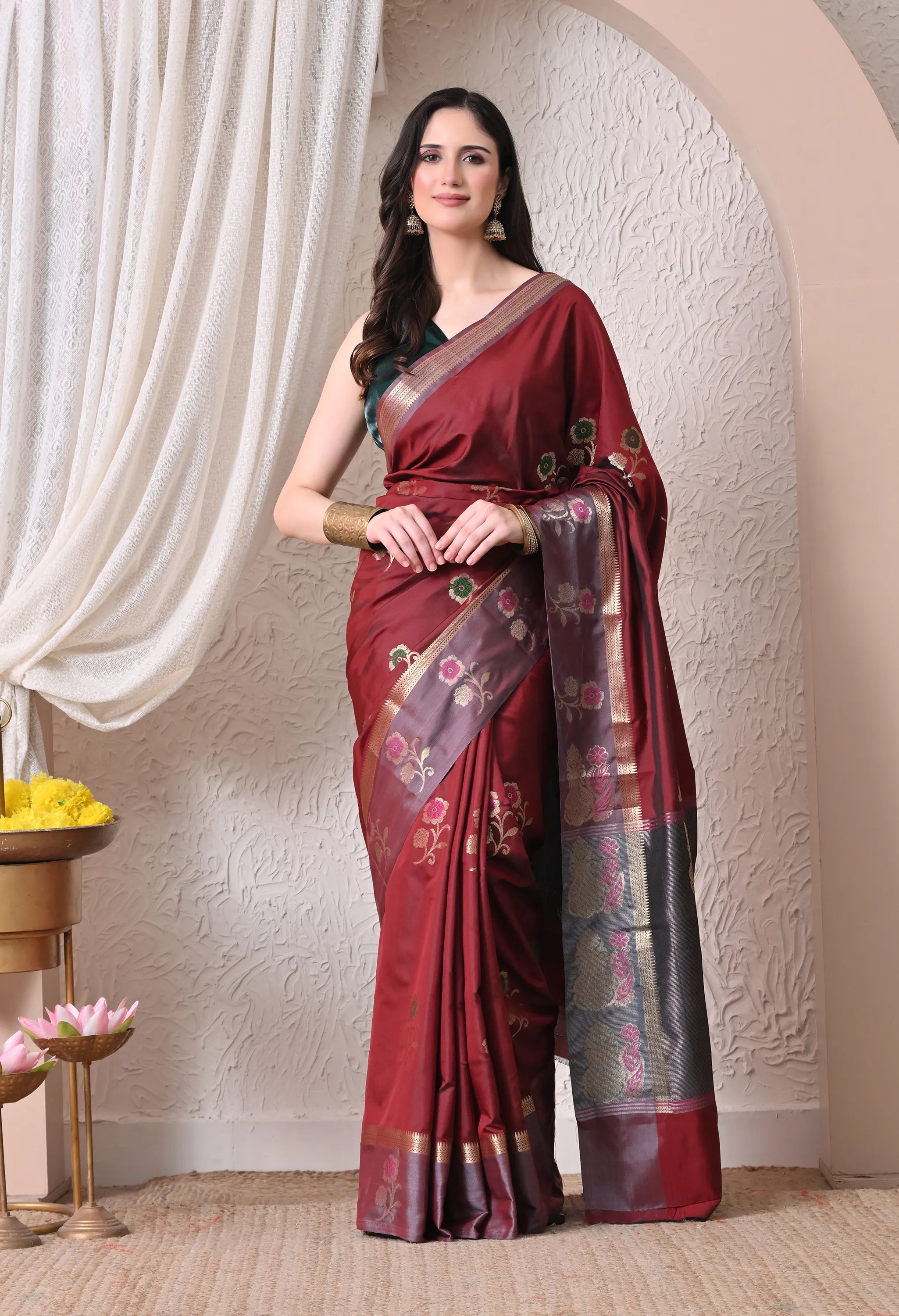 A woman poses in a rich maroon silk blend saree featuring a contrasting grey pallu and border adorned with delicate pink and green floral zari motifs.