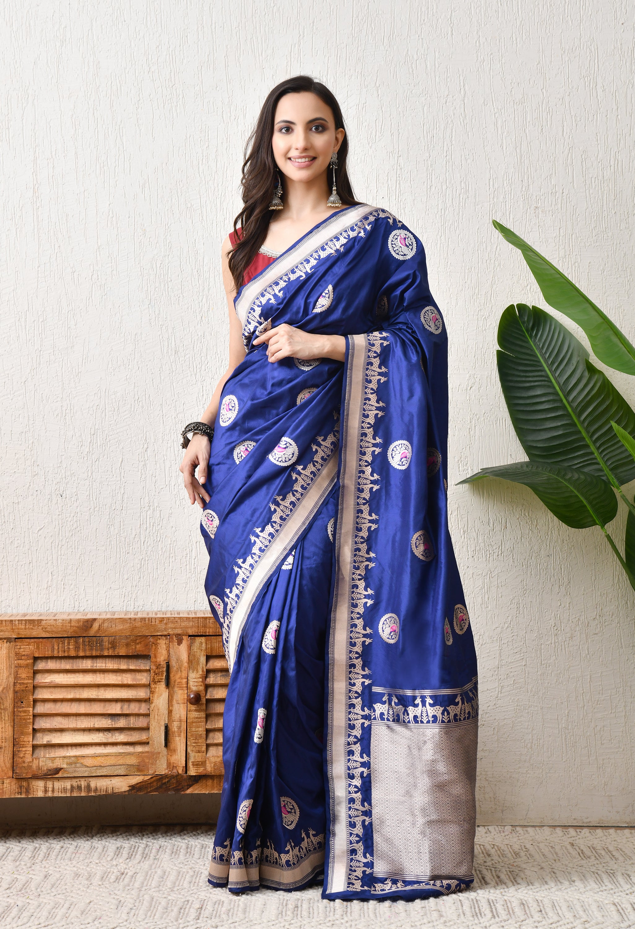 A woman modelling a royal blue Hand Woven Katan silk saree from The Weavers Shop by Raavi. The saree features golden zari circular motifs with bird designs across the body and a traditional Shikarga border depicting rows of deer. This Silk Mark Certified garment displays a rich lustre and is elegantly draped, paired with a maroon sleeveless blouse and traditional silver Jhumka earrings.