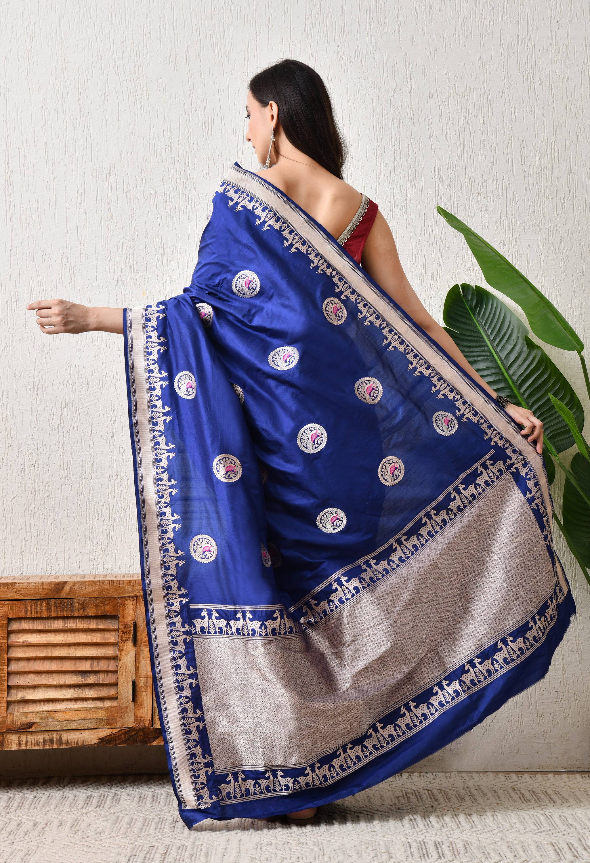 A back view of a woman showcasing the elegant drape and pallu of a royal blue Hand Woven Katan Silk saree by The Weavers Shop by Raavi. The image highlights the intricate golden zari Shikarga border featuring rows of deer and circular bird-design butis woven across the body. This Silk Mark Certified saree is paired with a maroon backless blouse, emphasizing the garment's rich luster and traditional craftsmanship.