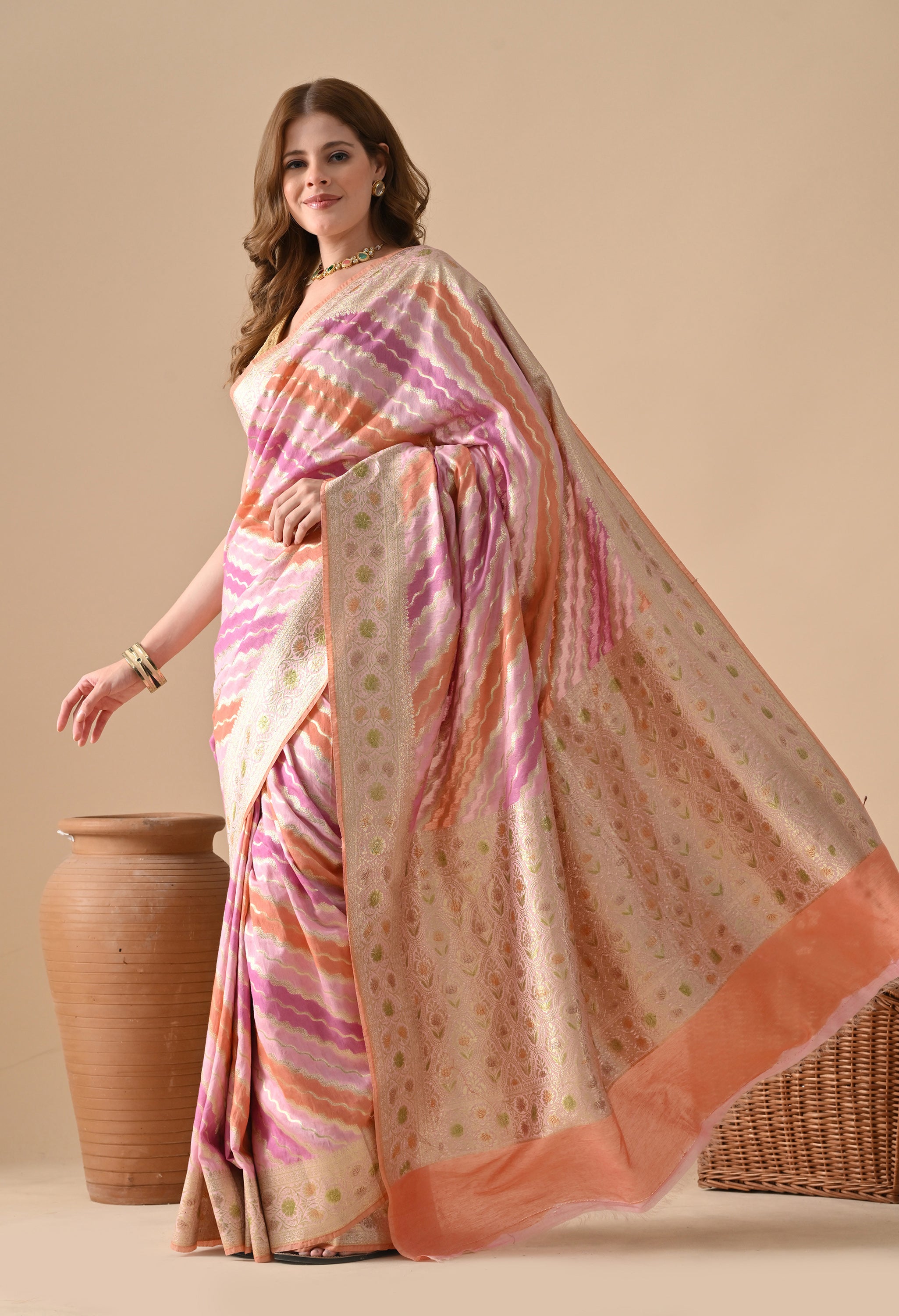A side profile of a woman modeling a multi-colored Banarasi silk saree by The Weavers Shop by Raavi. The shot captures the flowing texture of the silk and the vibrant pink, purple, and orange Rangkat wavy patterns. The wide gold zari border is visible in detail, running along the length of the saree, paired with a sleeveless gold blouse and ethnic jewelry.