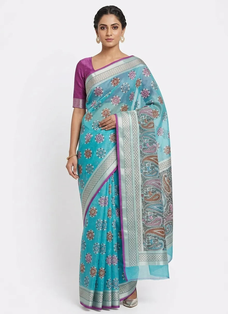A woman in a turquoise blue Banarasi silk blend saree featuring intricate red and white floral woven motifs. The saree has a contrasting silver zari border with purple piping and a grand pallu with traditional paisley designs.