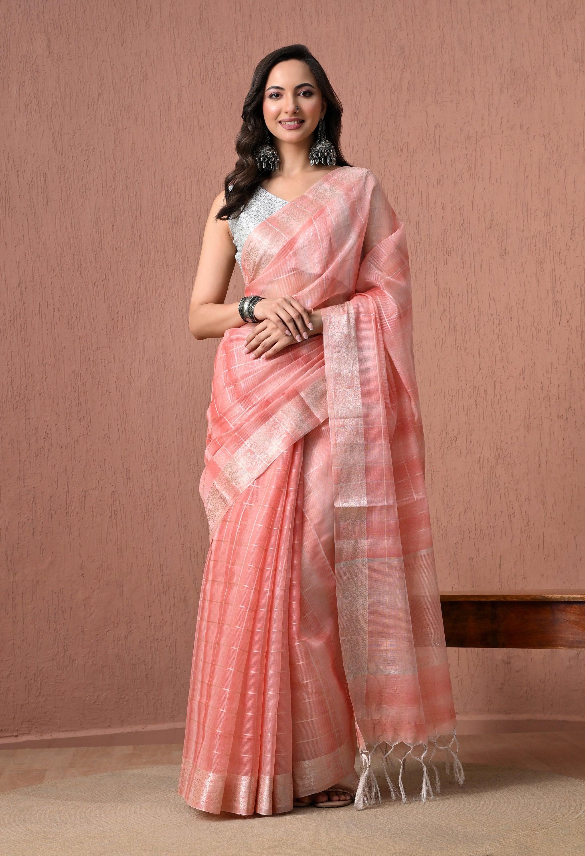 A full-length studio shot of a smiling woman modeling a light peach pure cotton saree with subtle check patterns and a silver zari border. She wears a sleeveless white textured blouse, large oxidized silver earrings, and matching bangles against a textured peach wall background.
