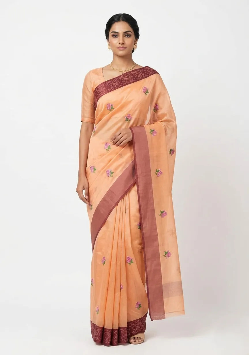 A woman posing in a peach silk blend Banarasi saree featuring small pink and green floral embroidered motifs. The saree has a contrasting maroon border and pallu with detailed woven designs and gold zari work. She is wearing a matching peach short-sleeved blouse and gold jewelry, set against a plain white background.