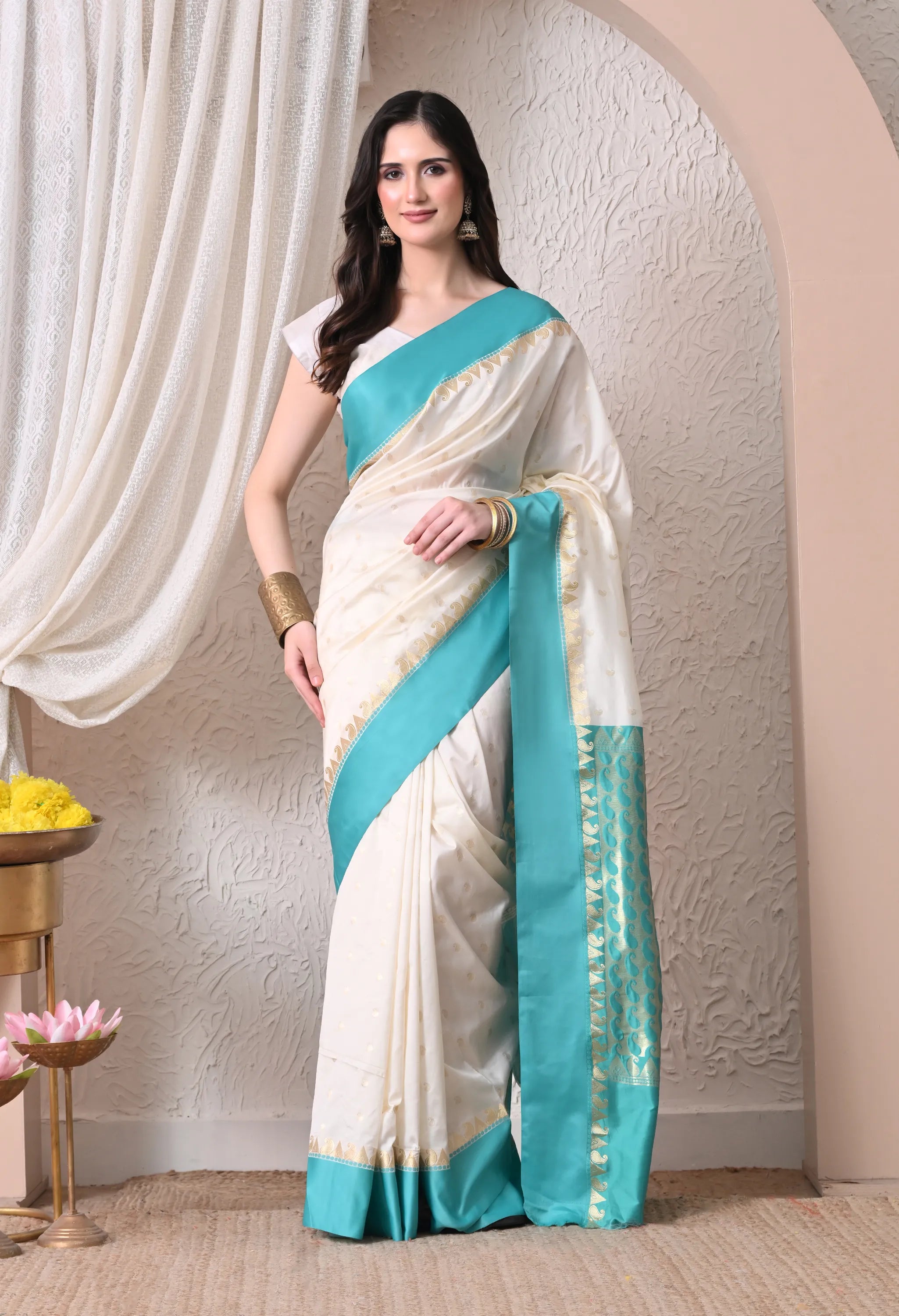 A woman showcasing an off-white silk blend saree accented by a vibrant turquoise blue border and pallu with golden zari details.
