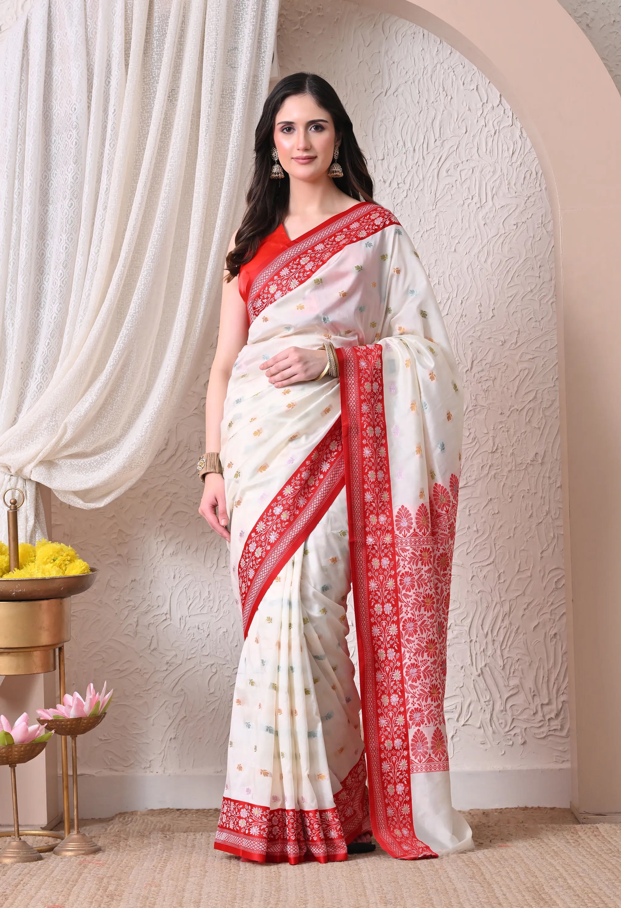 A woman drapes a traditional off-white silk blend saree featuring a rich red border and pallu with intricate woven patterns and small multi-colored body bootis
