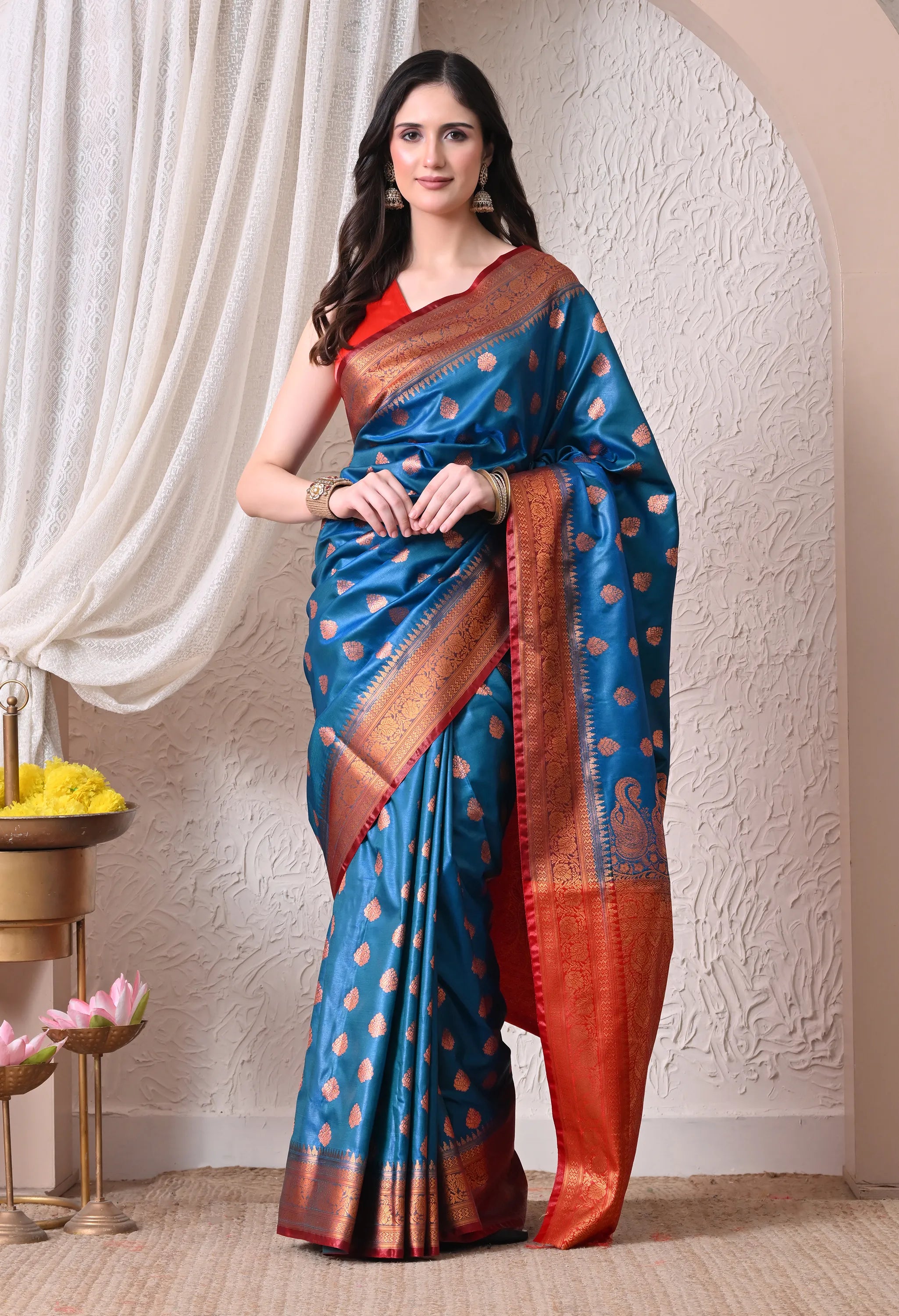 A woman standing elegantly in a peacock blue silk blend saree decorated with gold leaf motifs and a contrasting crimson red zari border and pallu.
