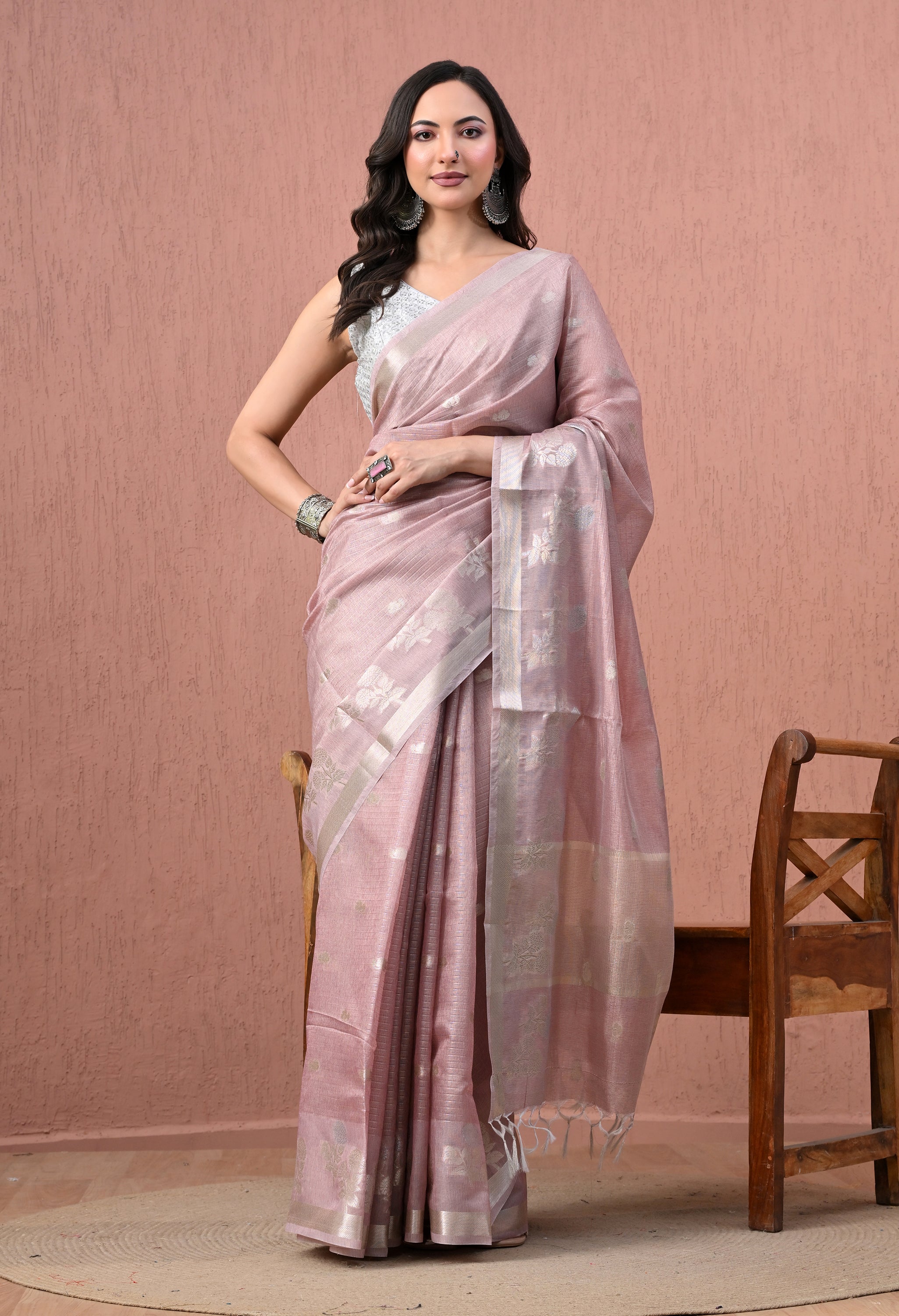 A full-length catalog photo of a model posing in a dusty rose pink pure cotton saree. The saree features woven silver zari floral patterns, a silver border, and tassel details on the pallu. She is wearing a sleeveless silver blouse, large oxidised silver earrings, a nose ring, and a heavy cuff bracelet, standing against a textured peach-pink wall.
