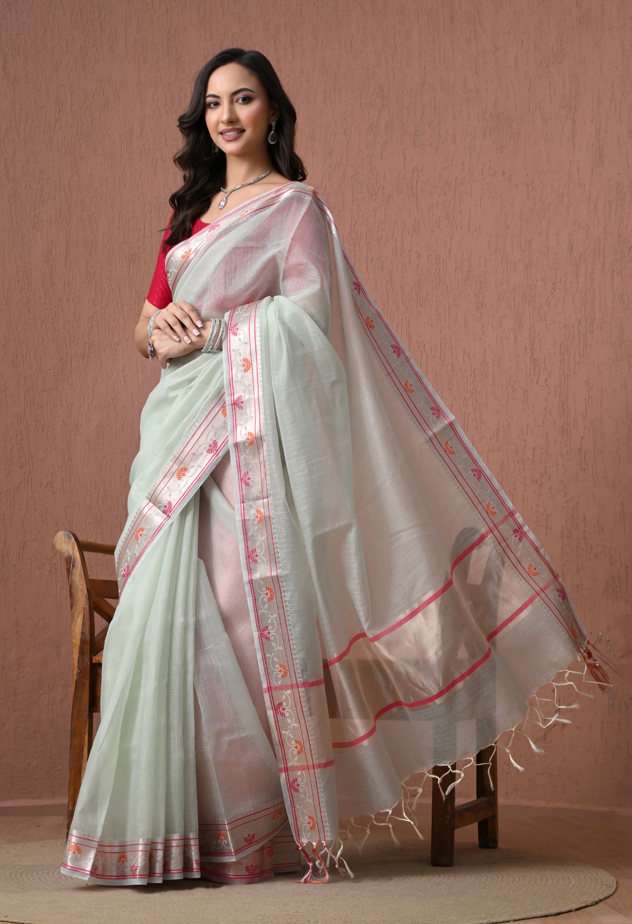 A woman showcasing a sheer pastel mint green pure cotton saree featuring a pink and orange floral border with silver accents, posed gracefully in front of a textured brown backdrop.
