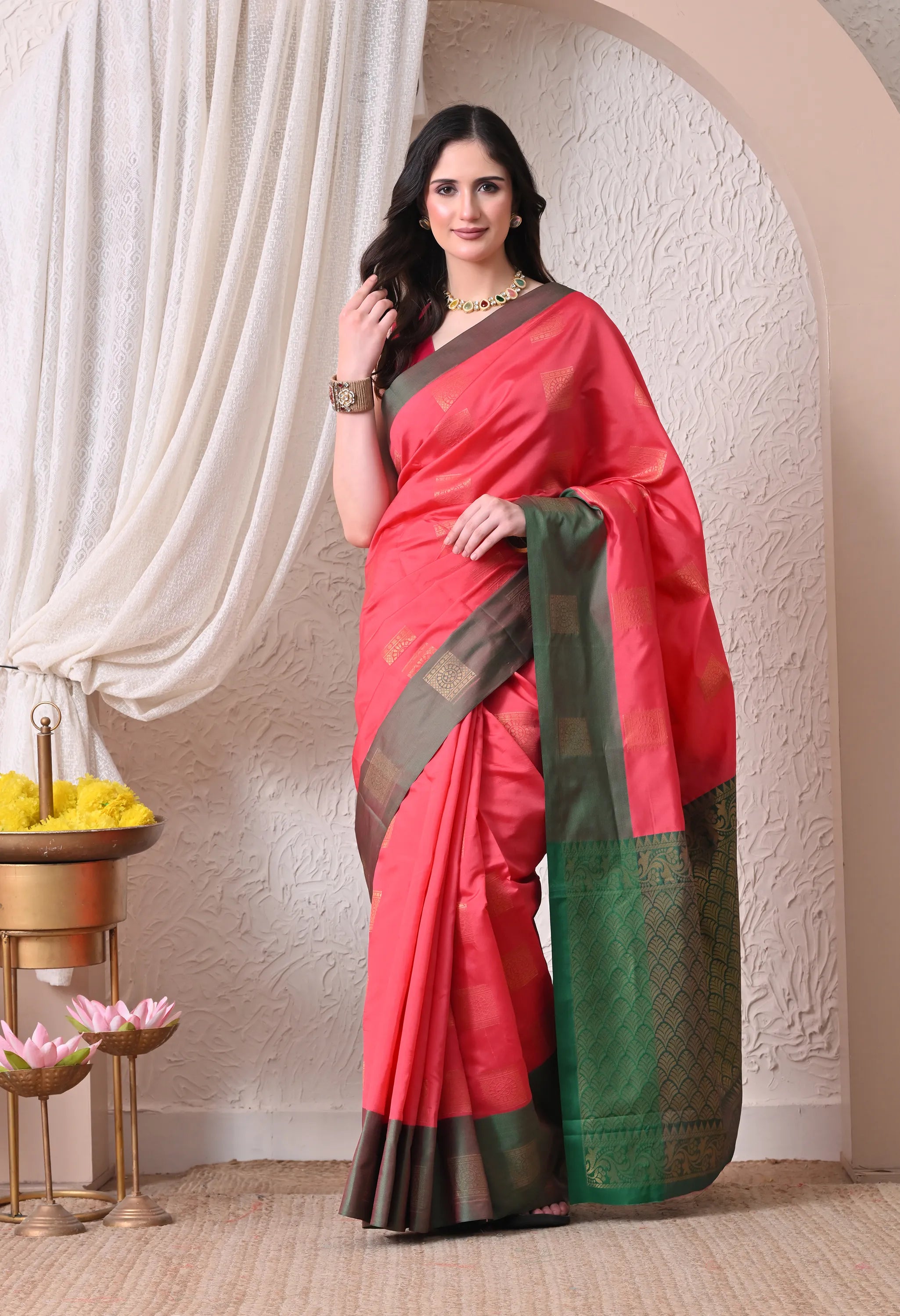 A woman posing in a vibrant coral pink silk blend saree featuring subtle square motifs and a contrasting deep olive green border and pallu.

