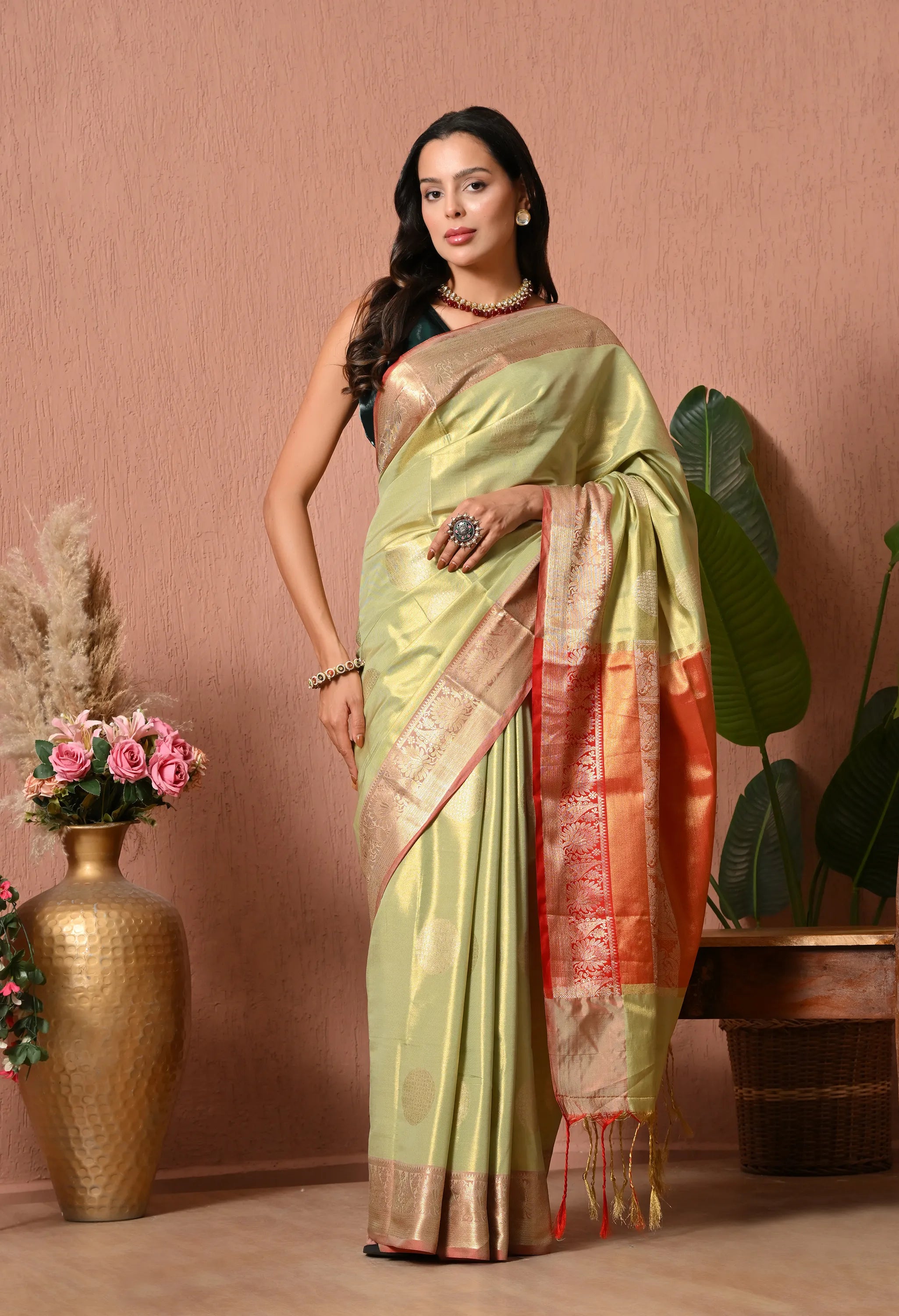 A woman gracefully carrying a pastel olive green silk blend saree adorned with circular gold motifs and a contrasting coral and copper zari border.
