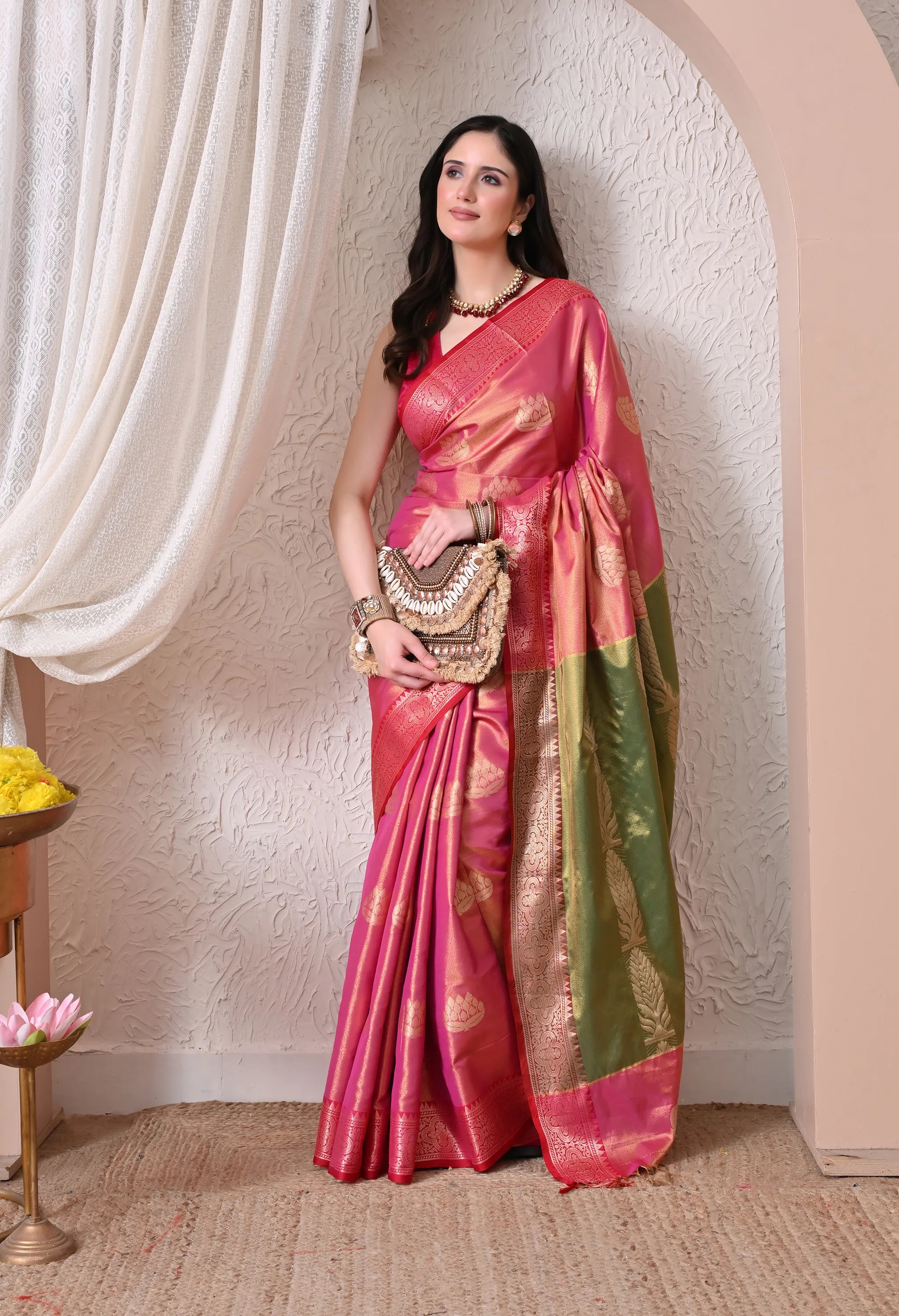 A woman posing elegantly in a Rani Pink silk blend saree with gold lotus motifs, featuring a striking contrast green and gold zari pallu.
