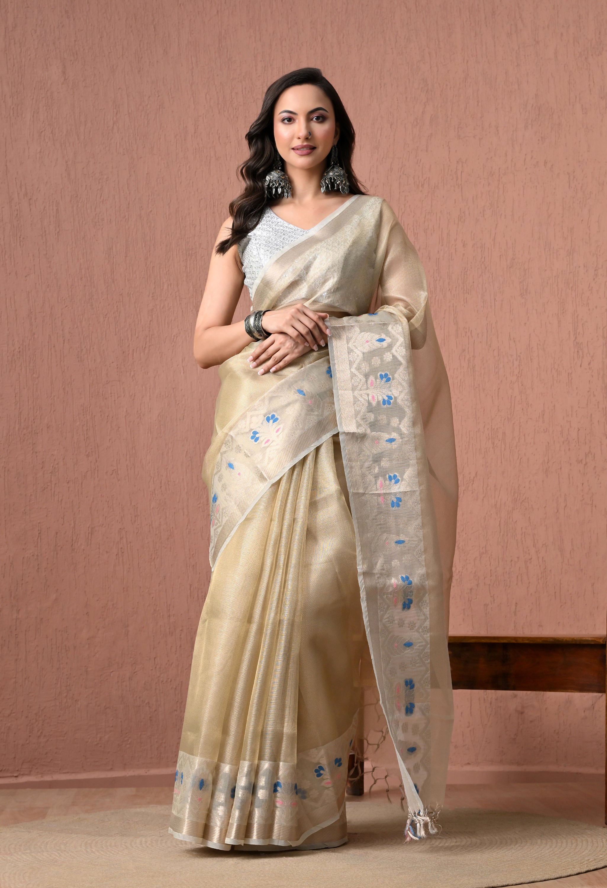 A full-length catalog photograph of a model gracefully posing in a metallic beige-gold pure cotton saree. The saree is adorned with a silver zari border and a heavily patterned pallu featuring small blue and pink woven floral details. She is wearing a sleeveless silver blouse, prominent oxidised silver jhumkas, and dark bangles against a textured dusty-rose background.
