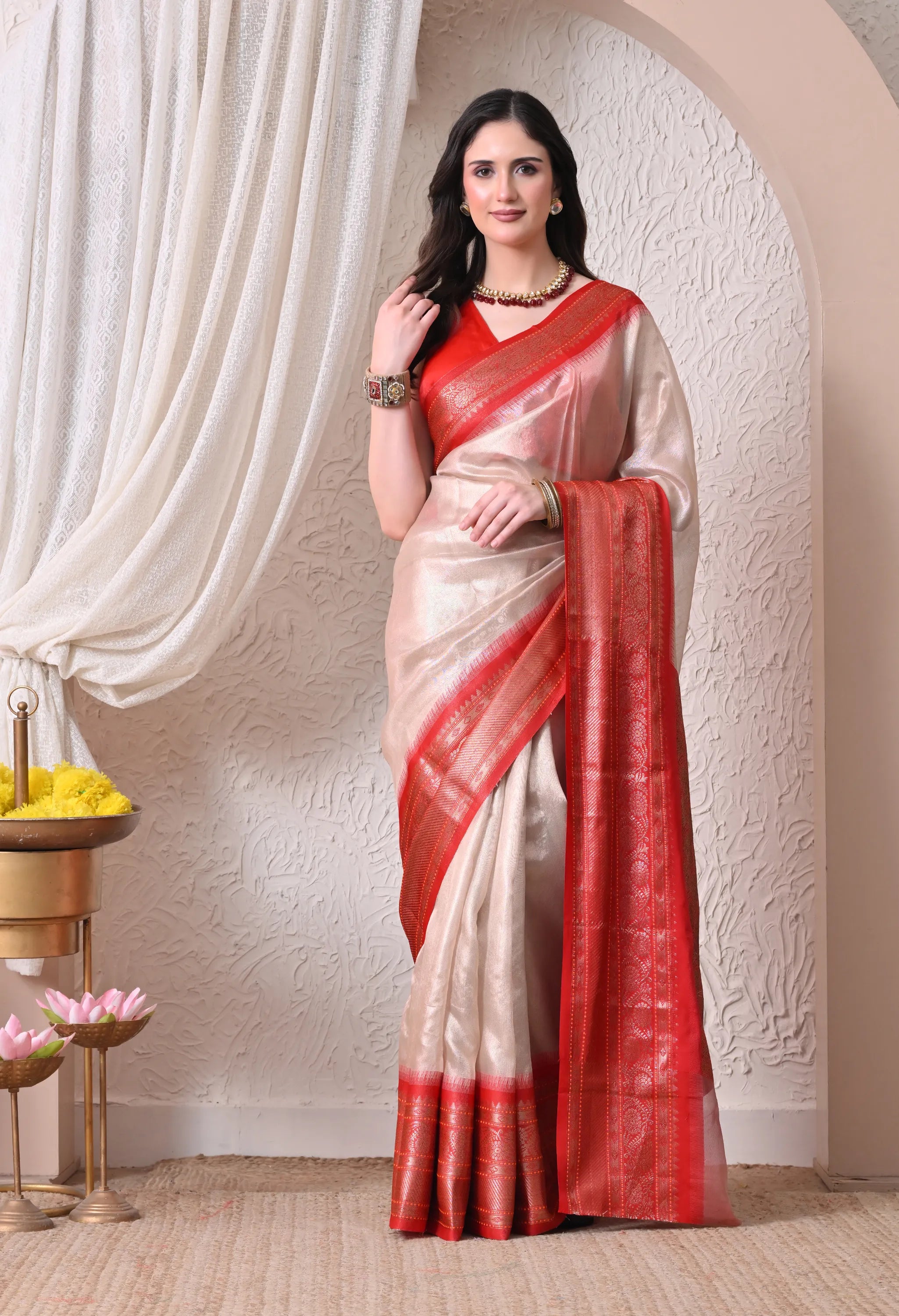 A woman gracefully posing in an ivory white tissue silk blend saree featuring a striking contrast vermilion red and gold zari border and pallu.
