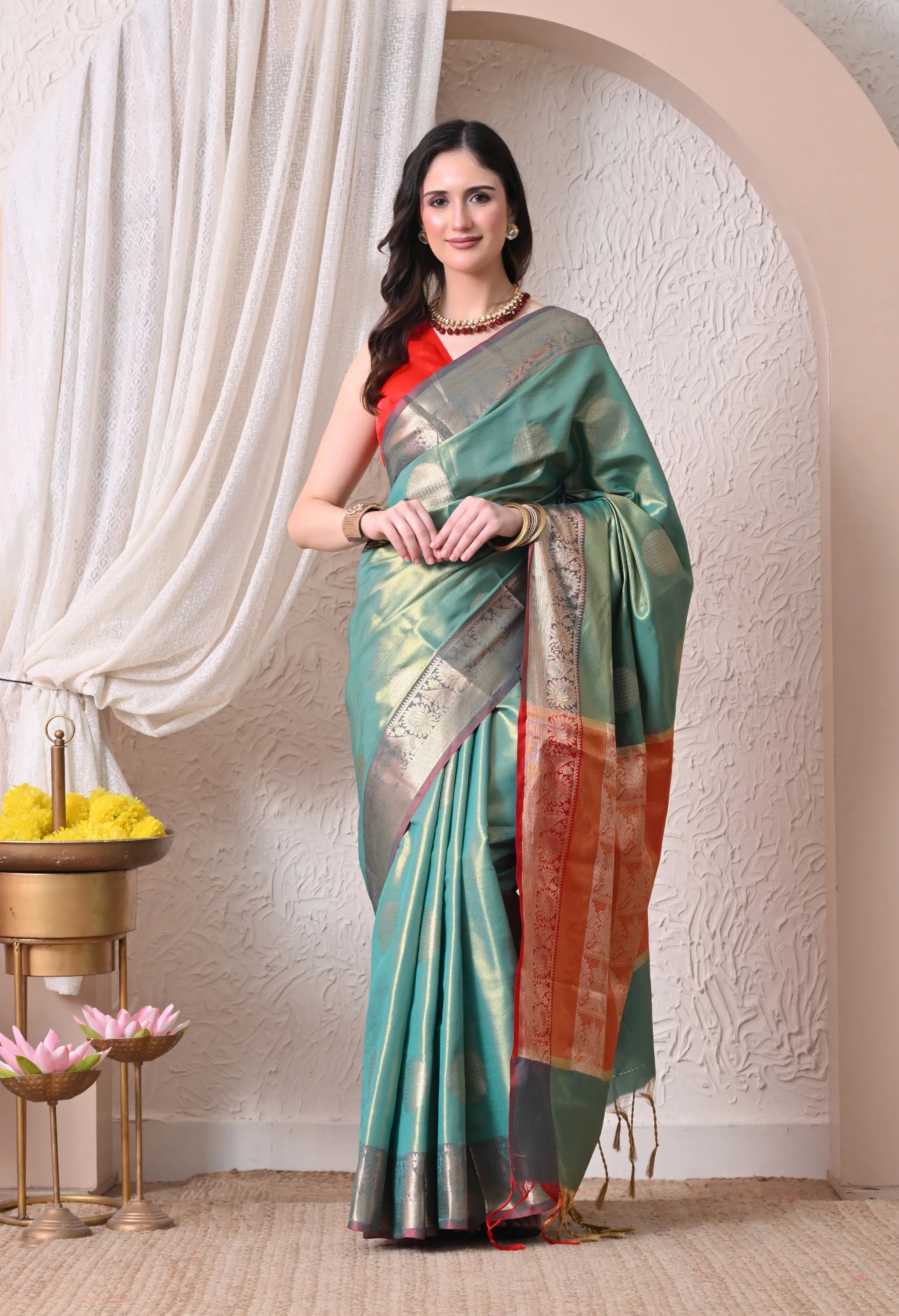 A woman gracefully wearing a seafoam green silk blend saree with large circular gold motifs and a contrasting coral red and gold zari border.
