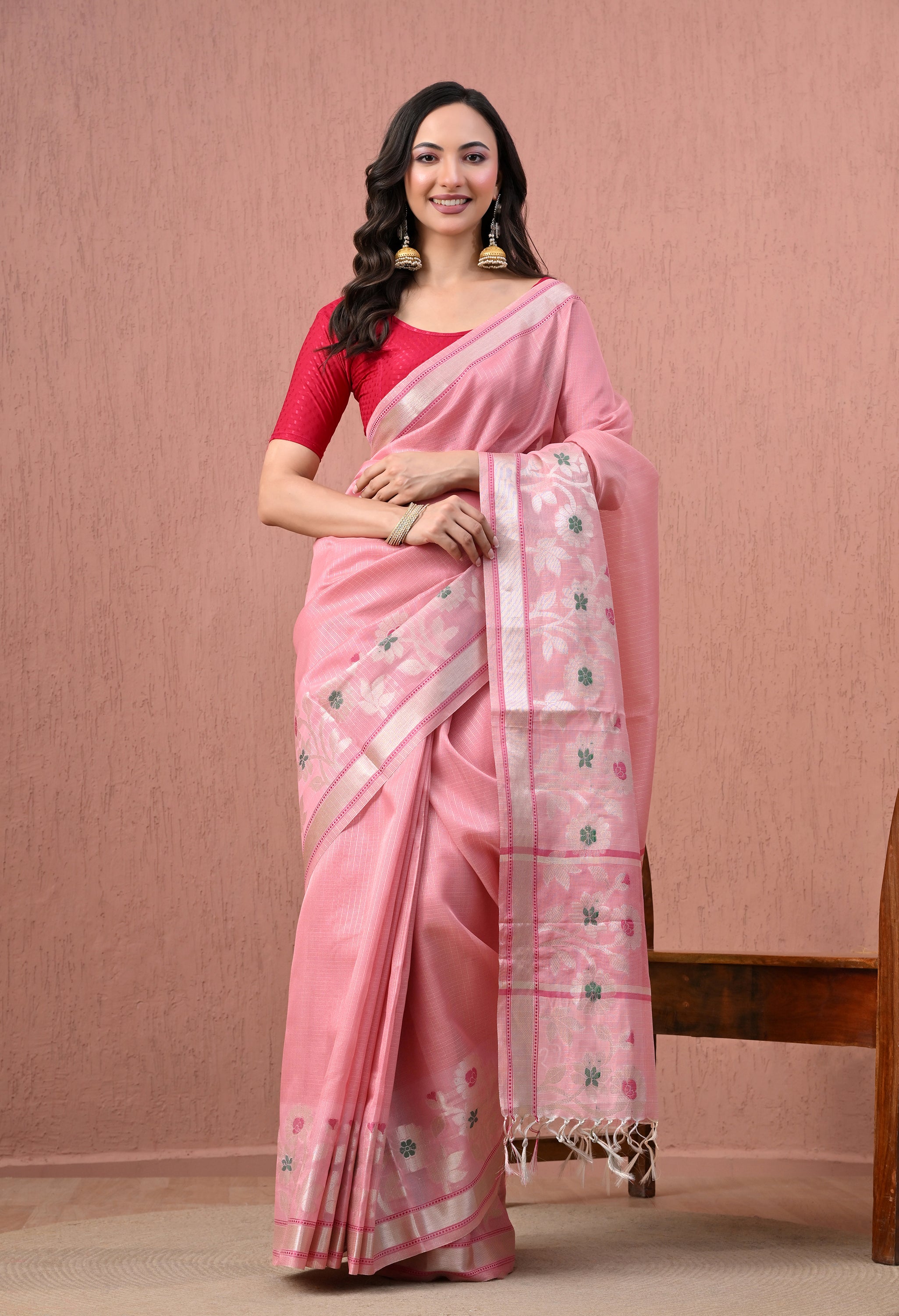 Full-length photograph of a woman smiling in a soft pink pure cotton handloom saree with a silver border and an intricately woven white and green floral pallu. She is wearing a contrasting crimson red blouse, traditional gold jhumka earrings, and standing against a textured dusty pink wall.
